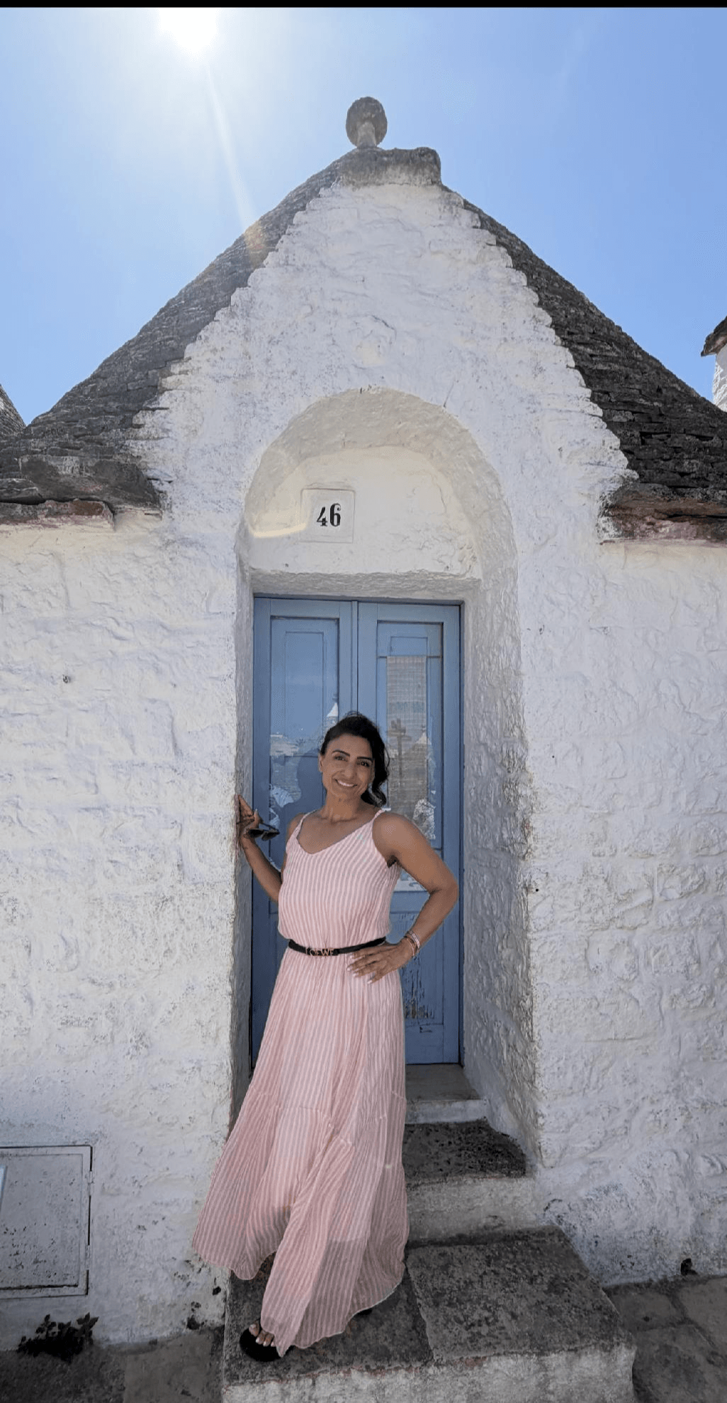 Woman in pink dress standing in front of a white building with a blue door, arched entrance, and a small bell on top.