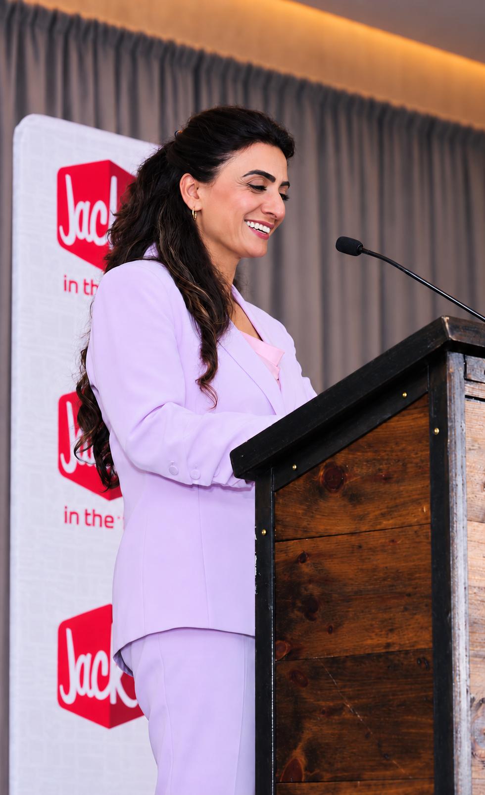 Woman with dark hair smiling at a podium during a presentation, with a Jack in the Box banner in the background.