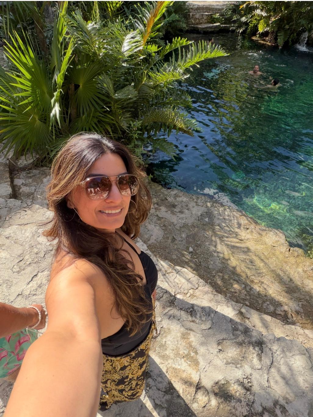 Woman taking selfie outdoors near water and lush green plants, wearing sunglasses and a black and yellow outfit.