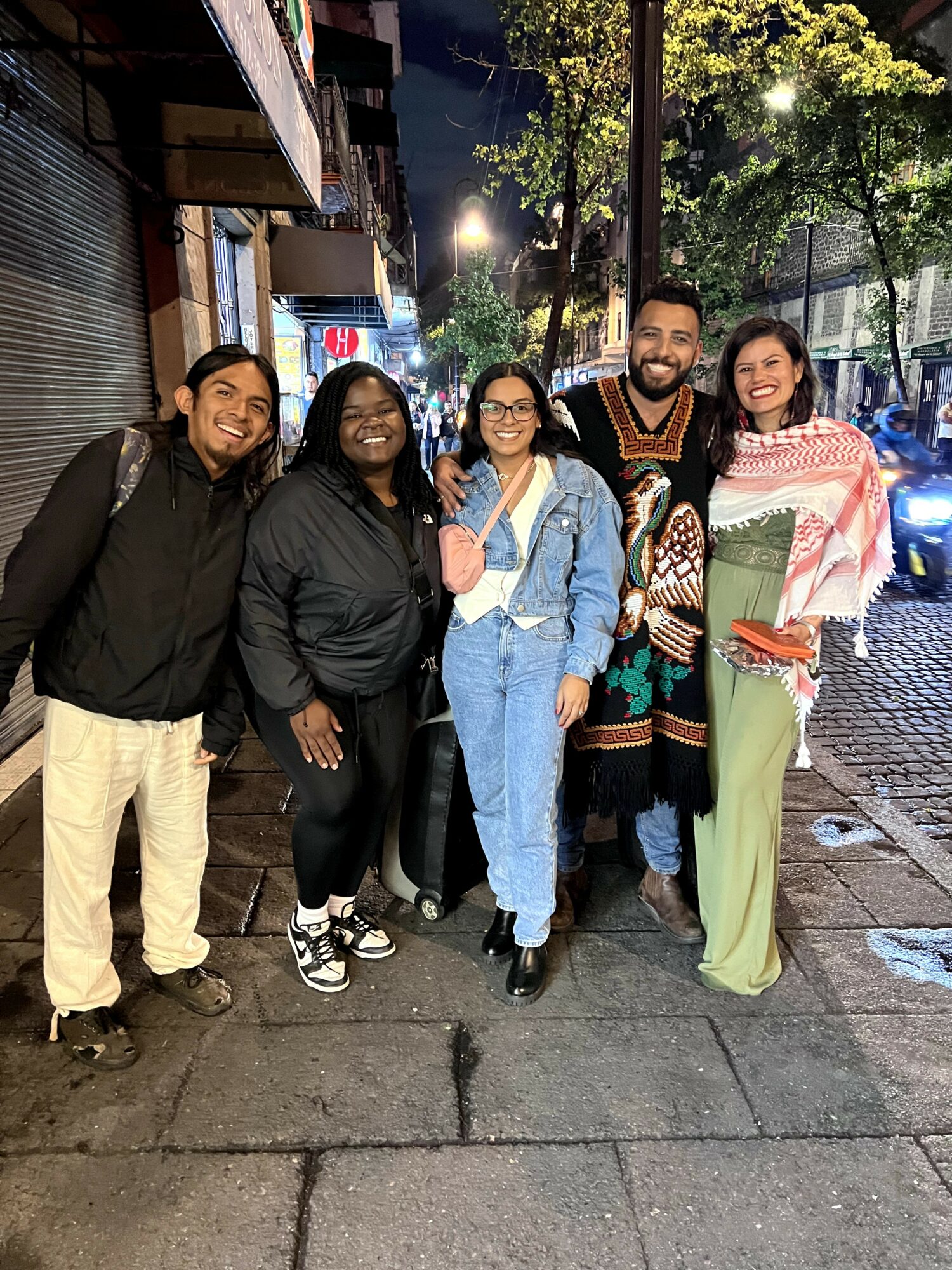 Group of five people standing on a sidewalk at night, smiling, with trees and streetlights in the background.
