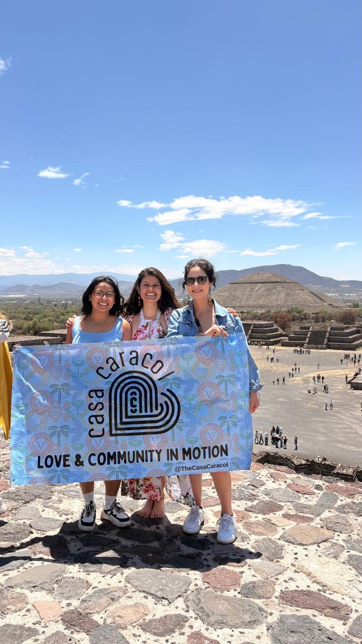 Three people holding a blue banner with a logo and text, standing outdoors on a stone surface with a historic site and mountains in the background.