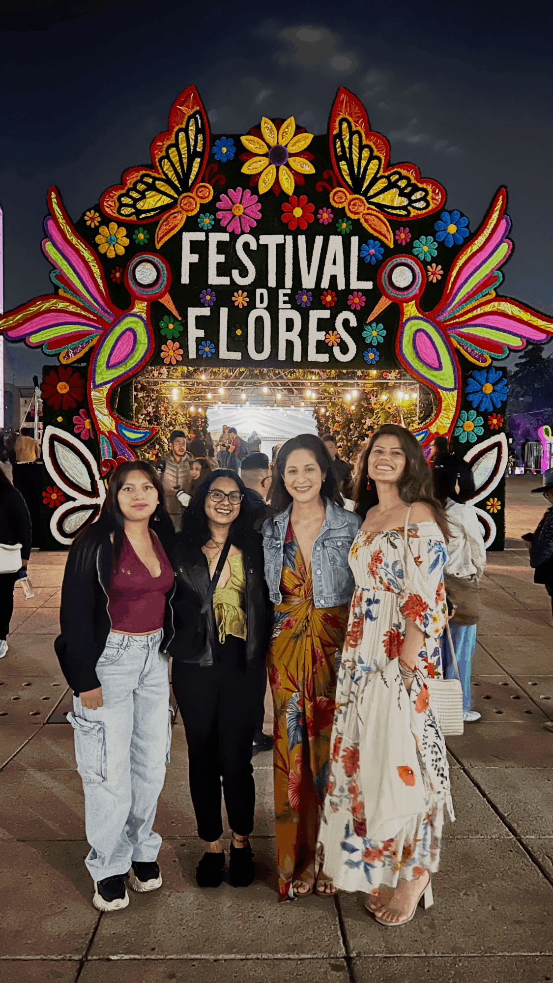 Four women standing in front of a colorful festival sign with flowers and butterflies, nighttime setting.