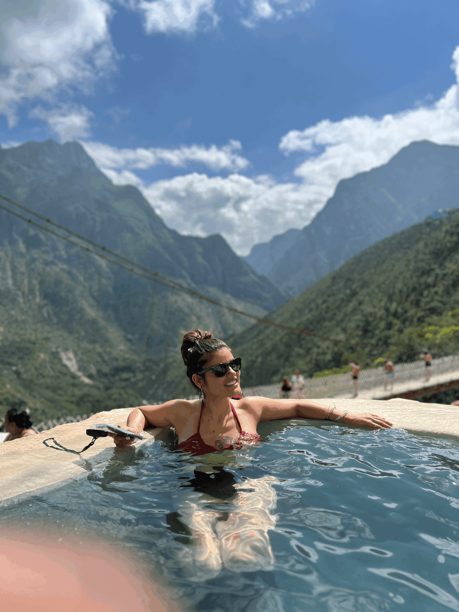Woman in sunglasses relaxing in a mountain pool with green mountains and blue sky in background.