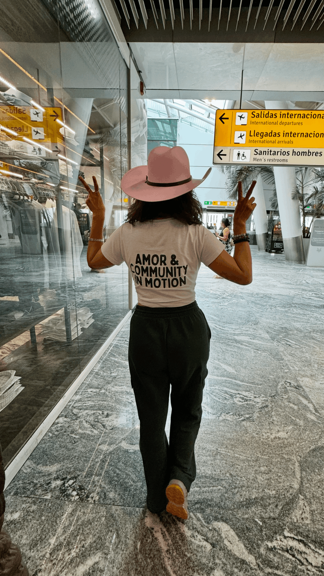 Person with pink hat and white shirt walking in an airport terminal, making peace signs, with signs in Spanish overhead.