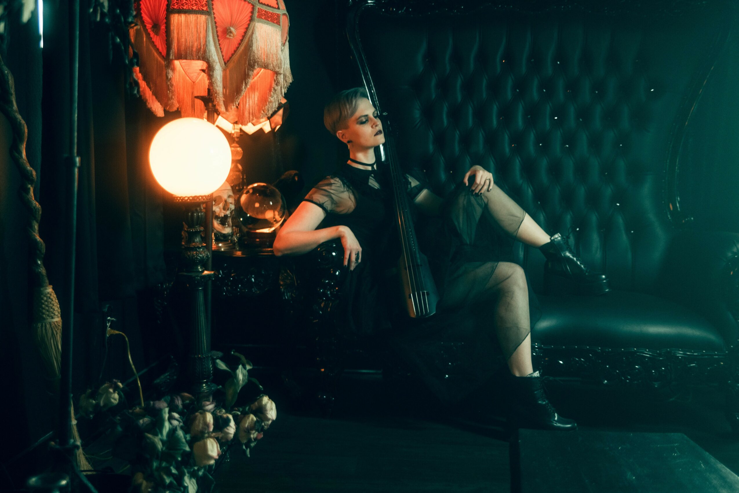 Person in black dress sitting on a black sofa with a cello, in a dimly lit room with a lamp and dark curtains.