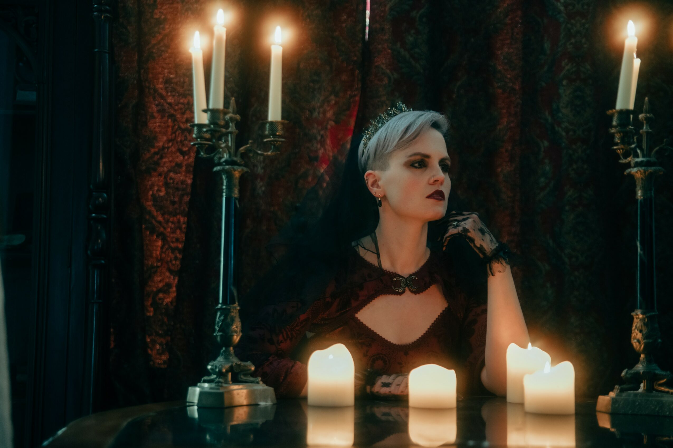 Young woman with short hair and dark makeup in a dark room with candles and ornate curtains.