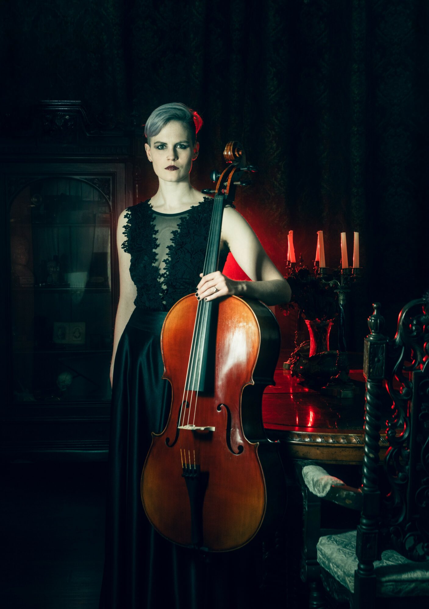 Woman in black dress holding a cello in a dimly lit room with dark curtains and vintage furniture.