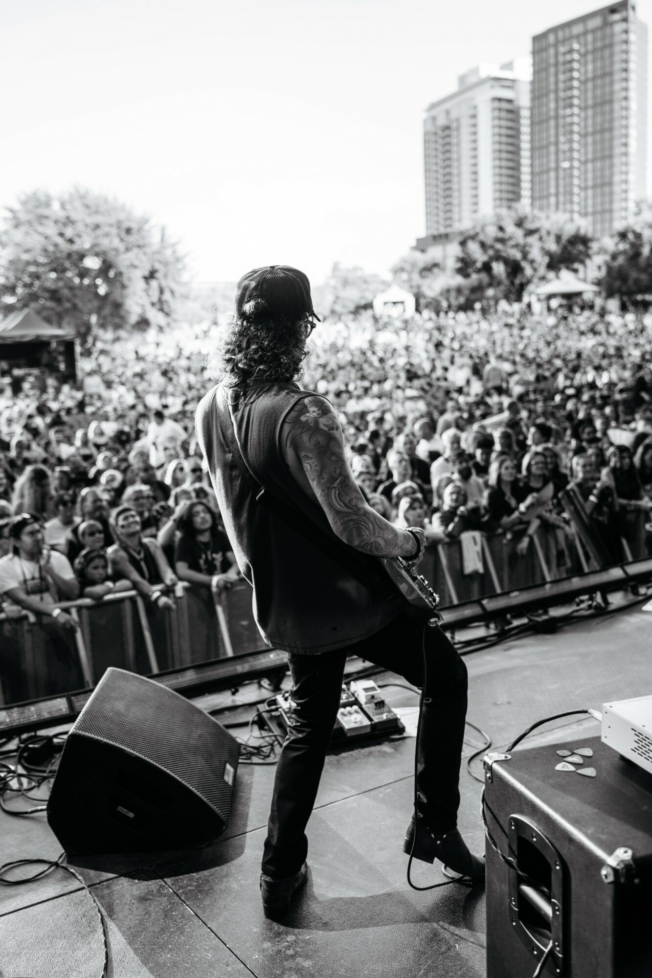 Musician performing on stage with a crowd and city buildings in background, black and white, from behind.