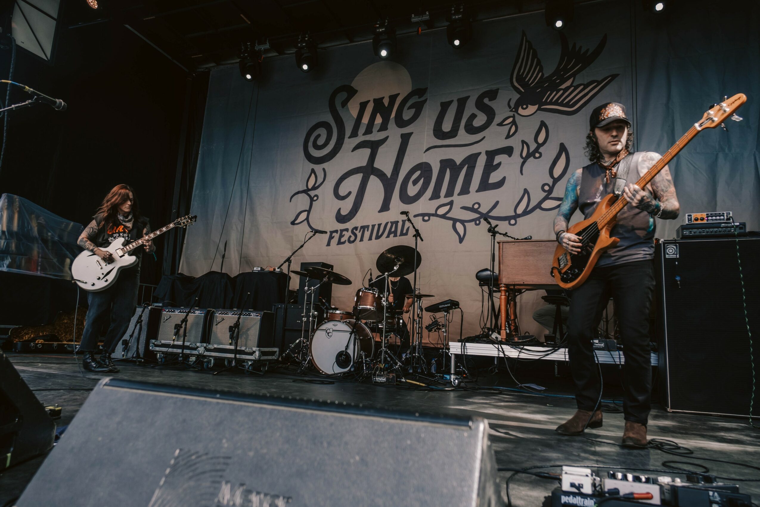 Band performing on stage at Sing Us Home Festival with drums, guitar, and bass, and a large festival banner in the background.