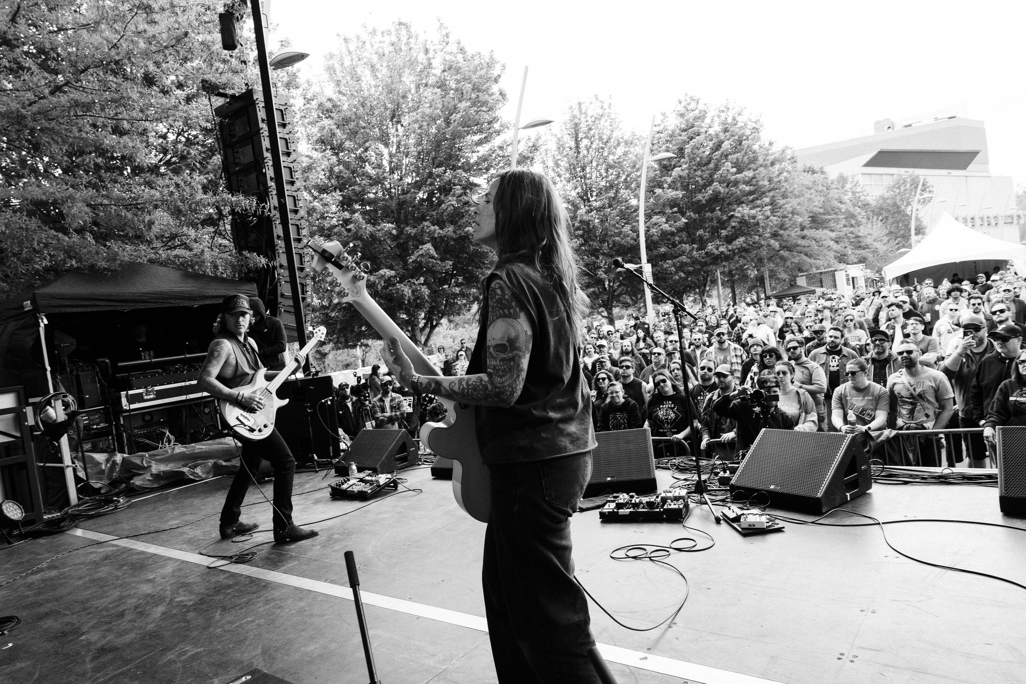 Musicians perform on stage outdoors with a crowd watching, trees and buildings in the background, black and white photo.