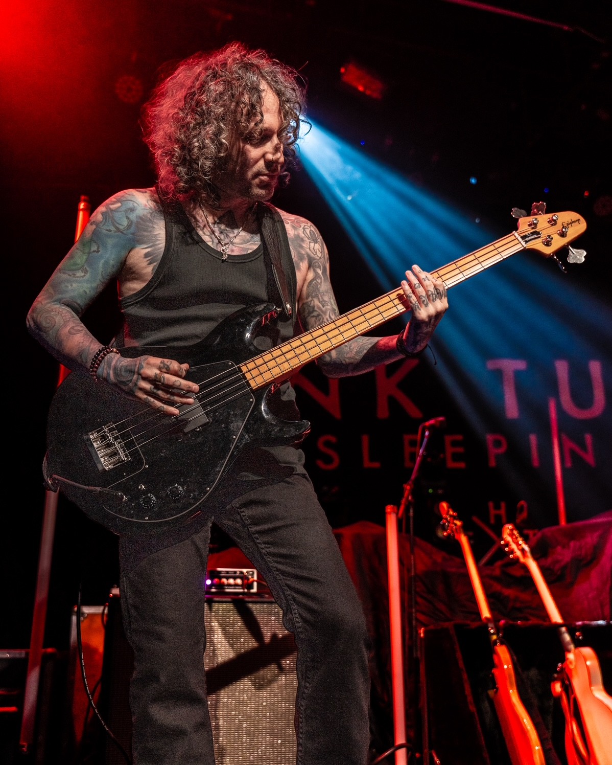 Person with curly hair playing a black electric bass guitar on stage, illuminated by blue and red lights.