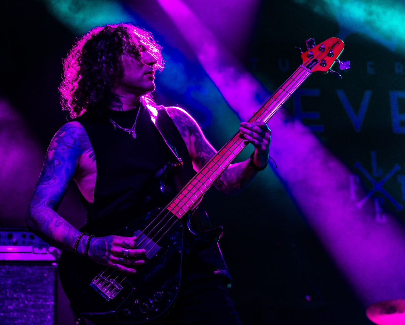 Person playing bass guitar on stage with colorful lighting, curly hair, sleeveless top, and tattoos.
