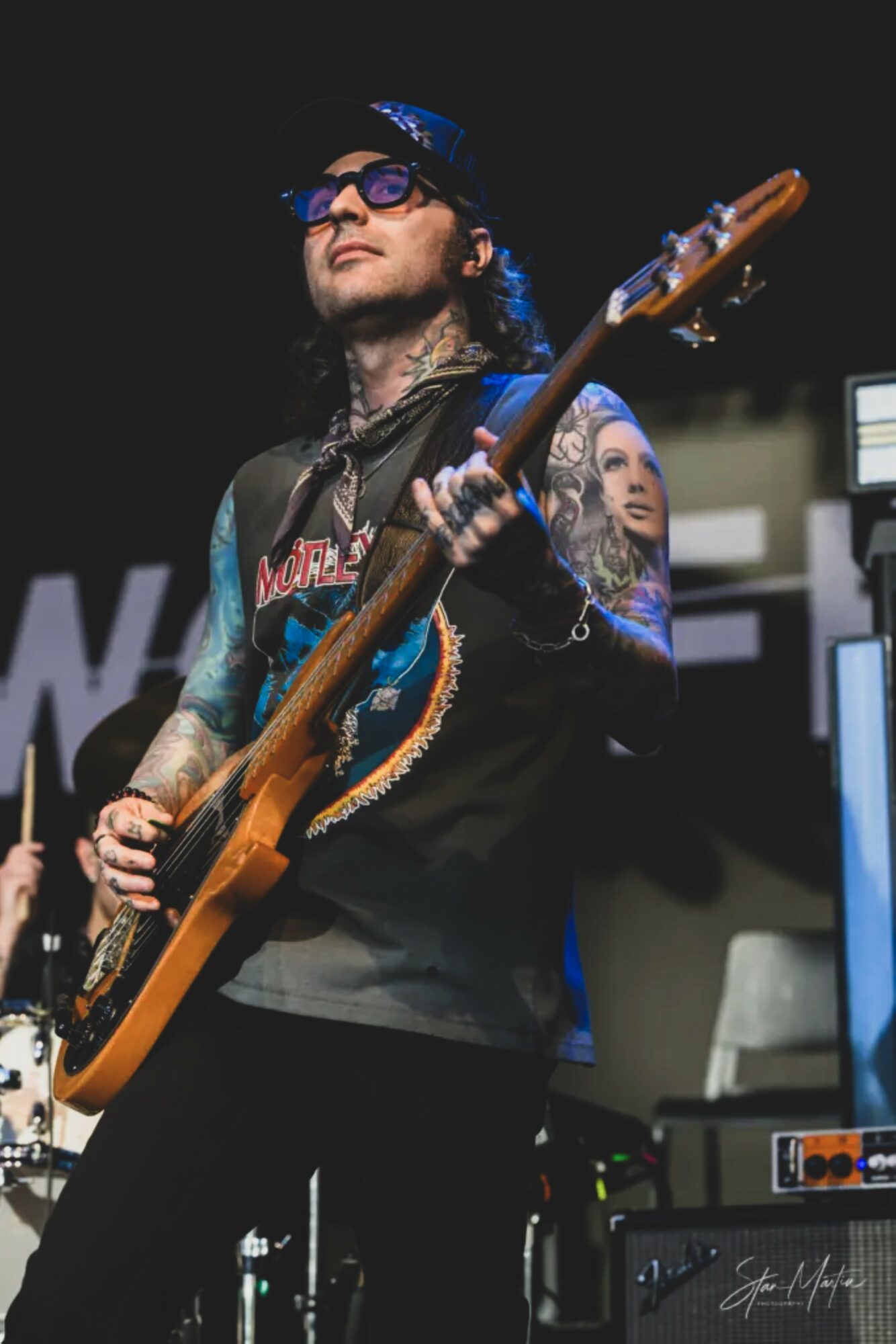Musician playing guitar on stage with dark background, wearing sunglasses, cap, and tattoos visible on arms.