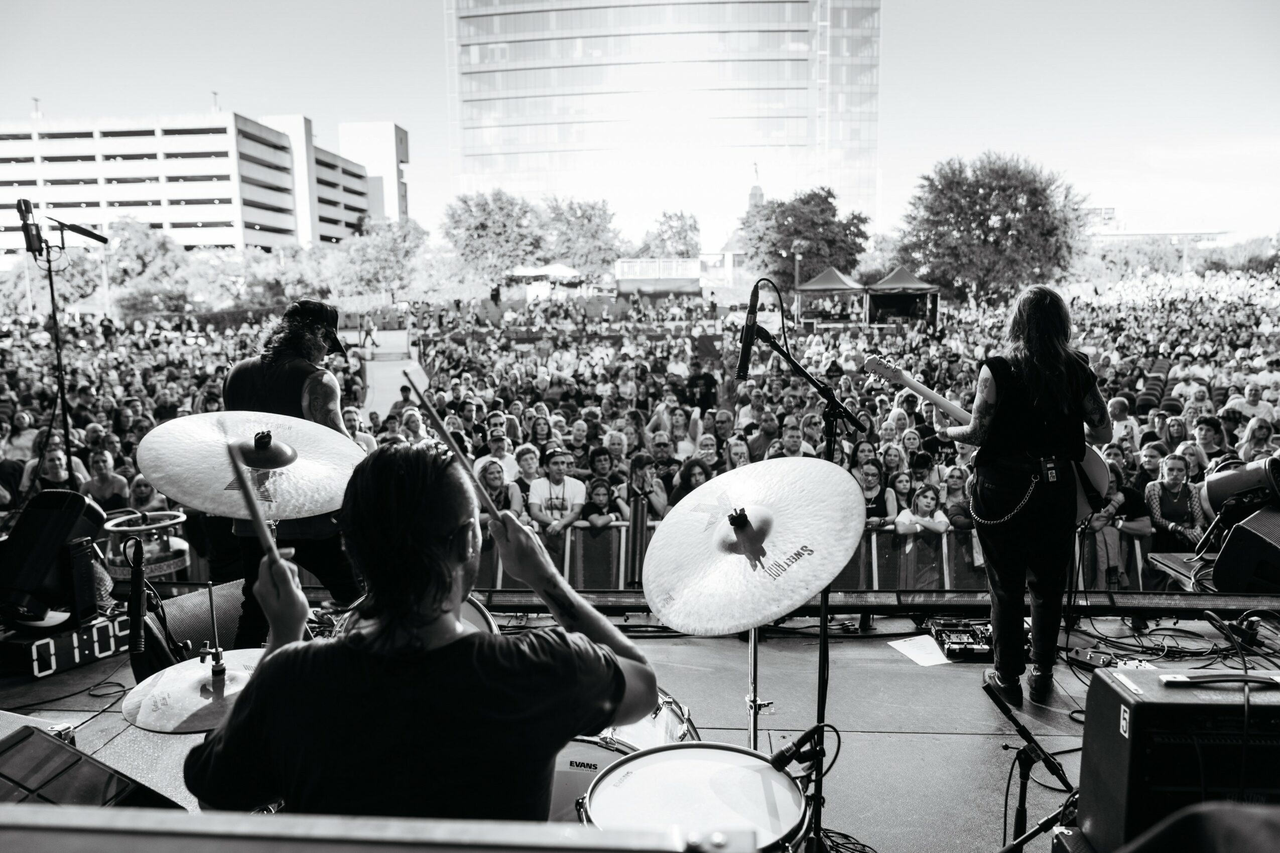 Band performs on stage in front of a large crowd outdoors, with buildings and trees in the background.