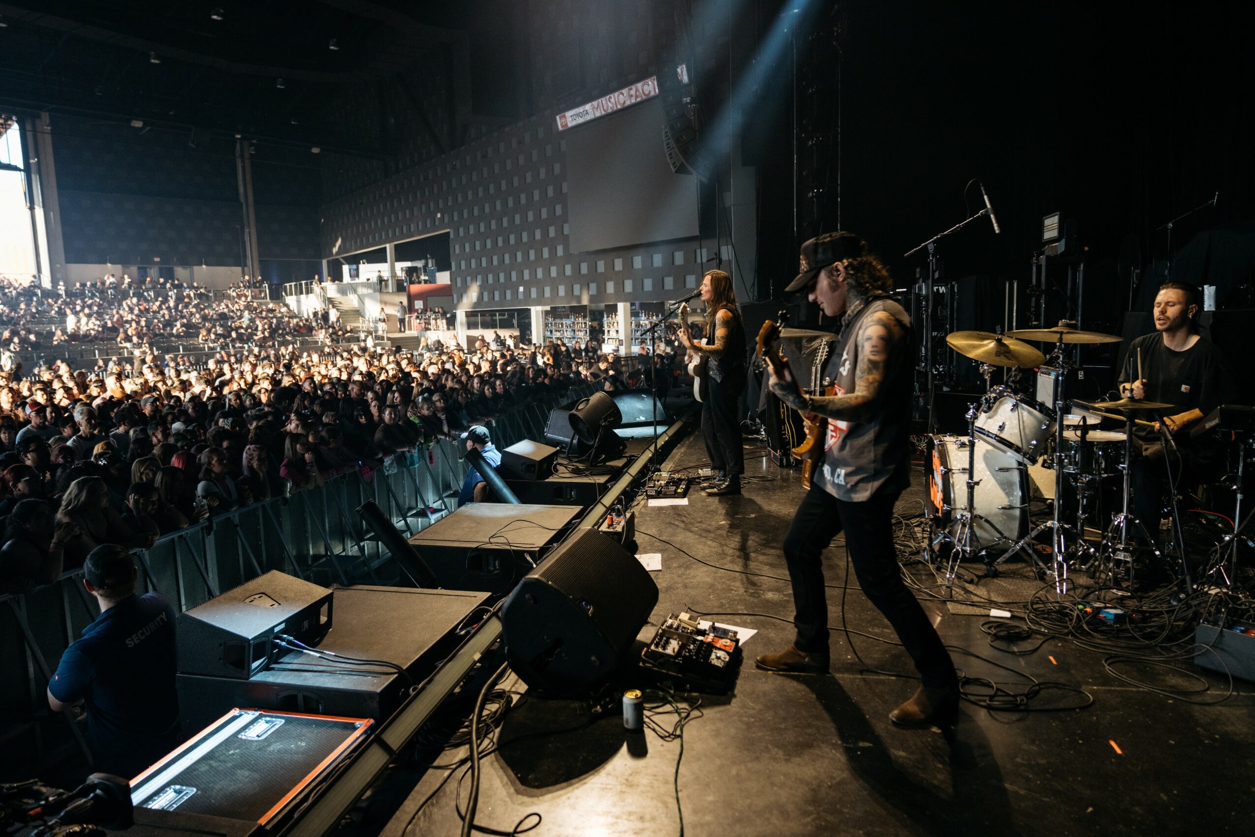 Band performs on stage with a large audience in a dark concert venue, stage lights shining down, musicians playing instruments.