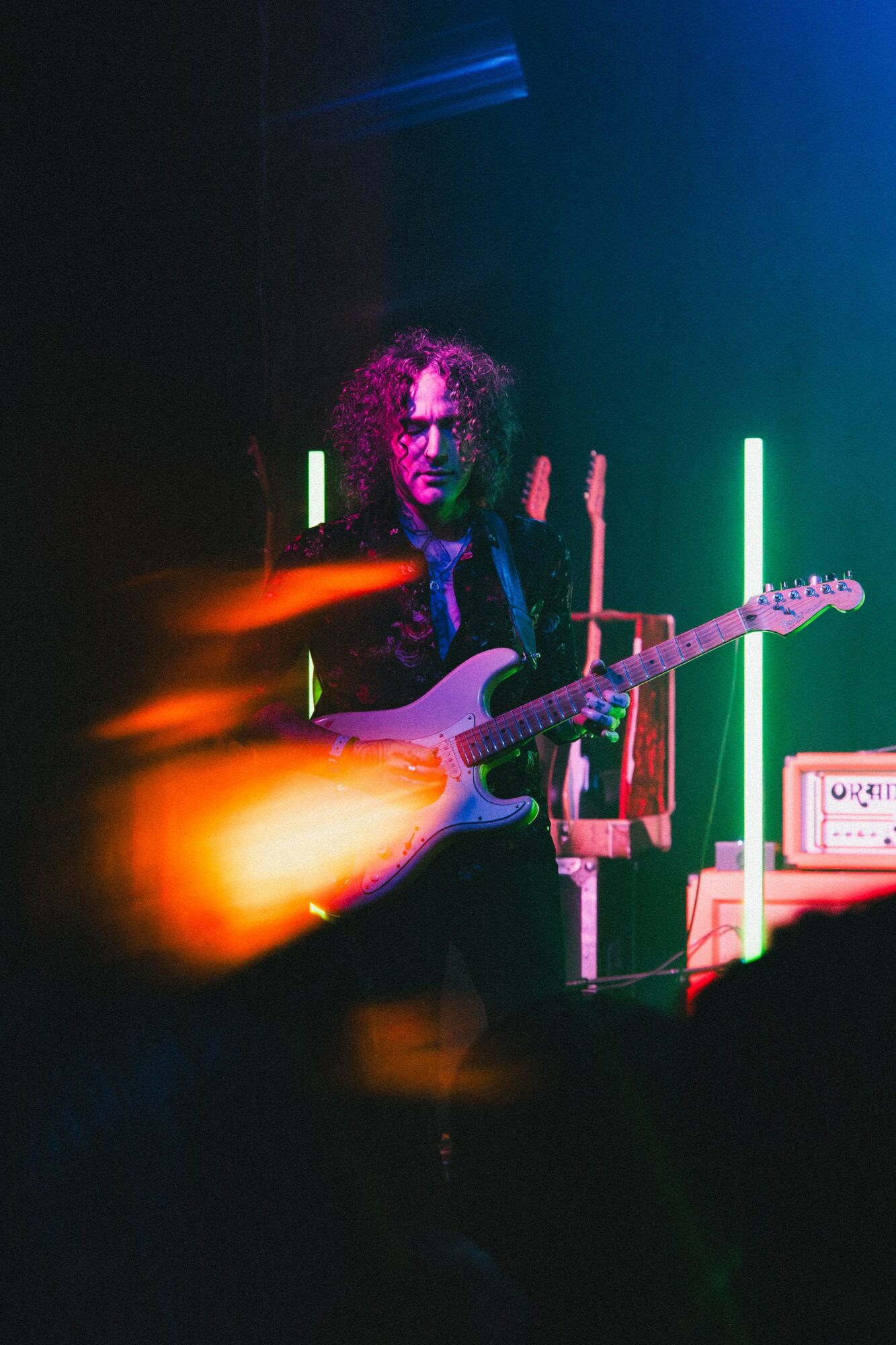 Person playing an electric guitar on stage with colorful vertical lights in the background.