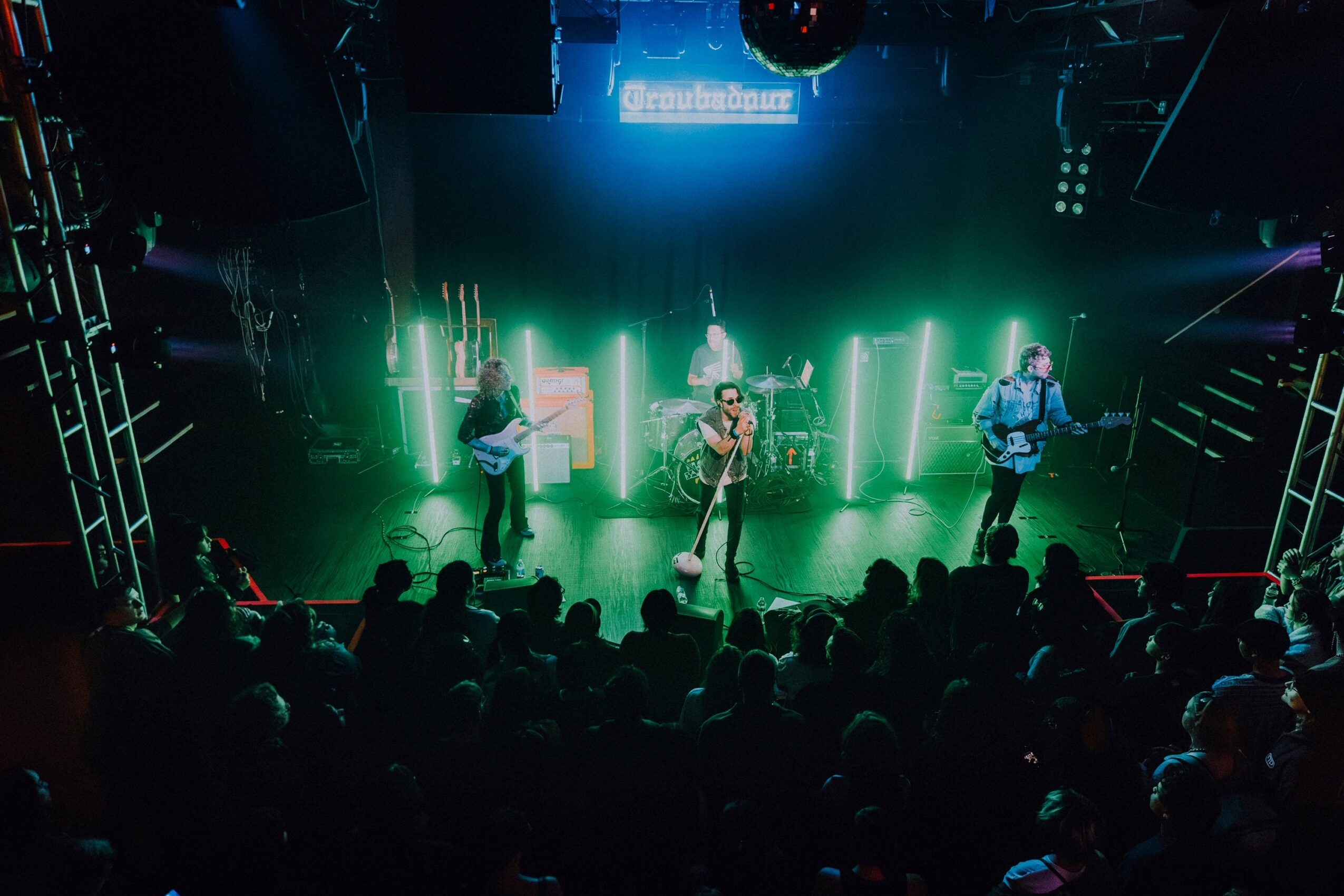Band performs on stage with colorful lights, audience watching, and a screen above displaying text.