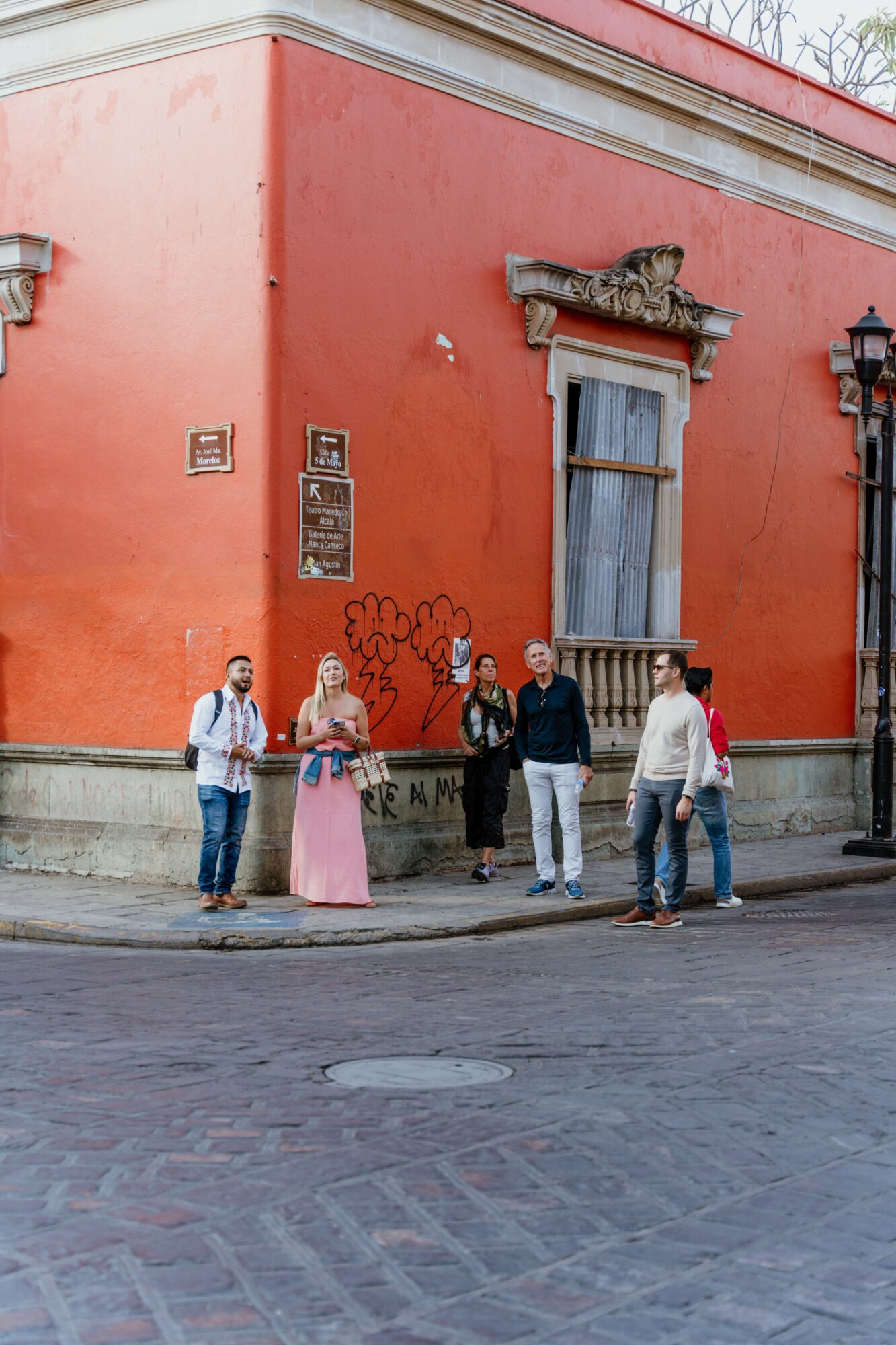 Group of five people standing on a sidewalk in front of a red building with a window and graffiti, street lamp nearby.