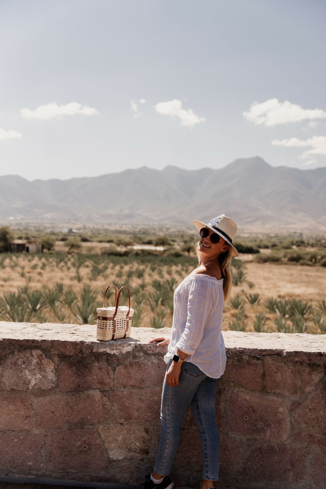 Woman wearing sunglasses and a hat stands by a stone wall with a desert landscape and mountains in the background.