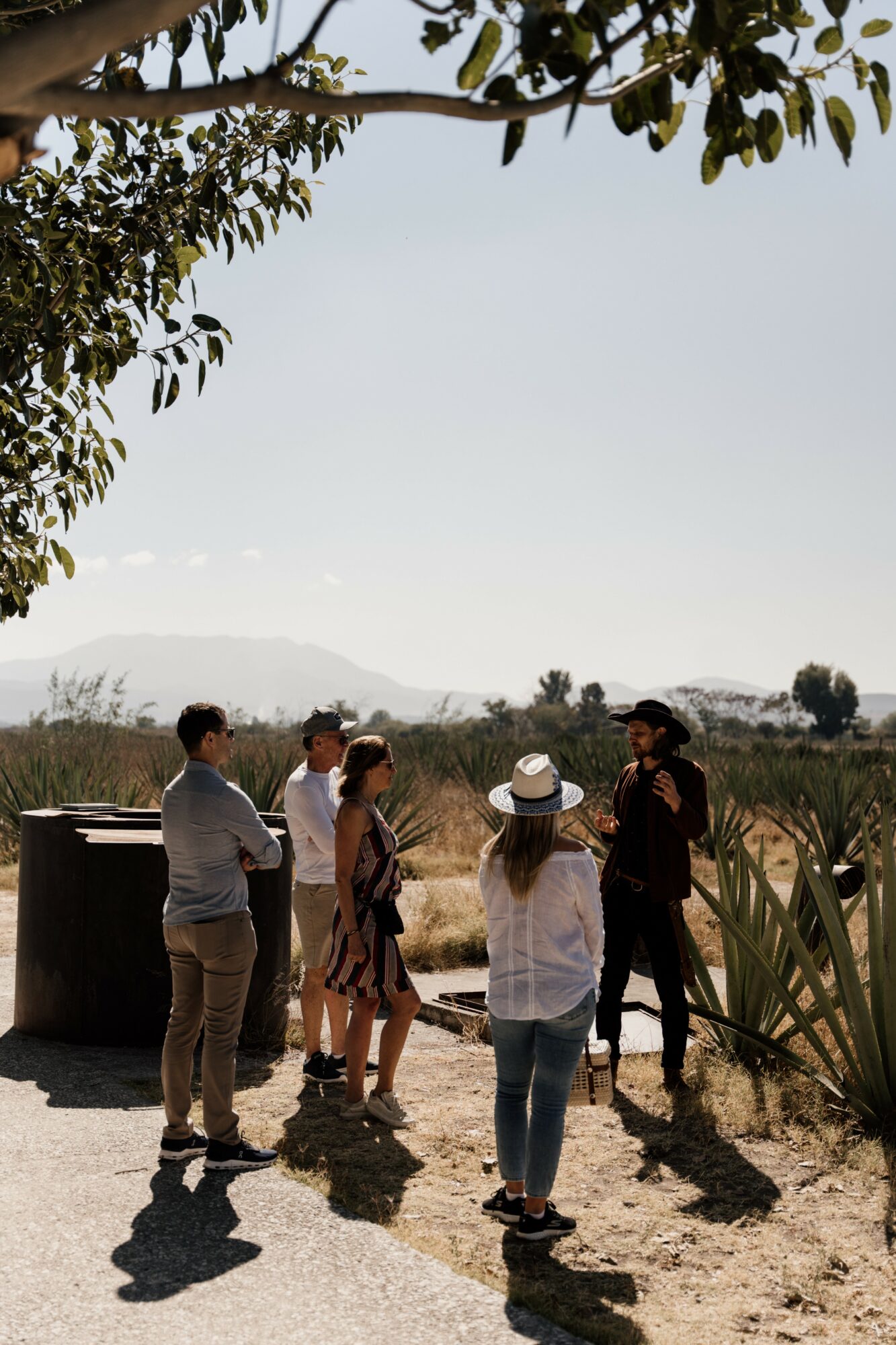 Group of six people standing outdoors near plants and trees, with a clear sky and distant landscape.