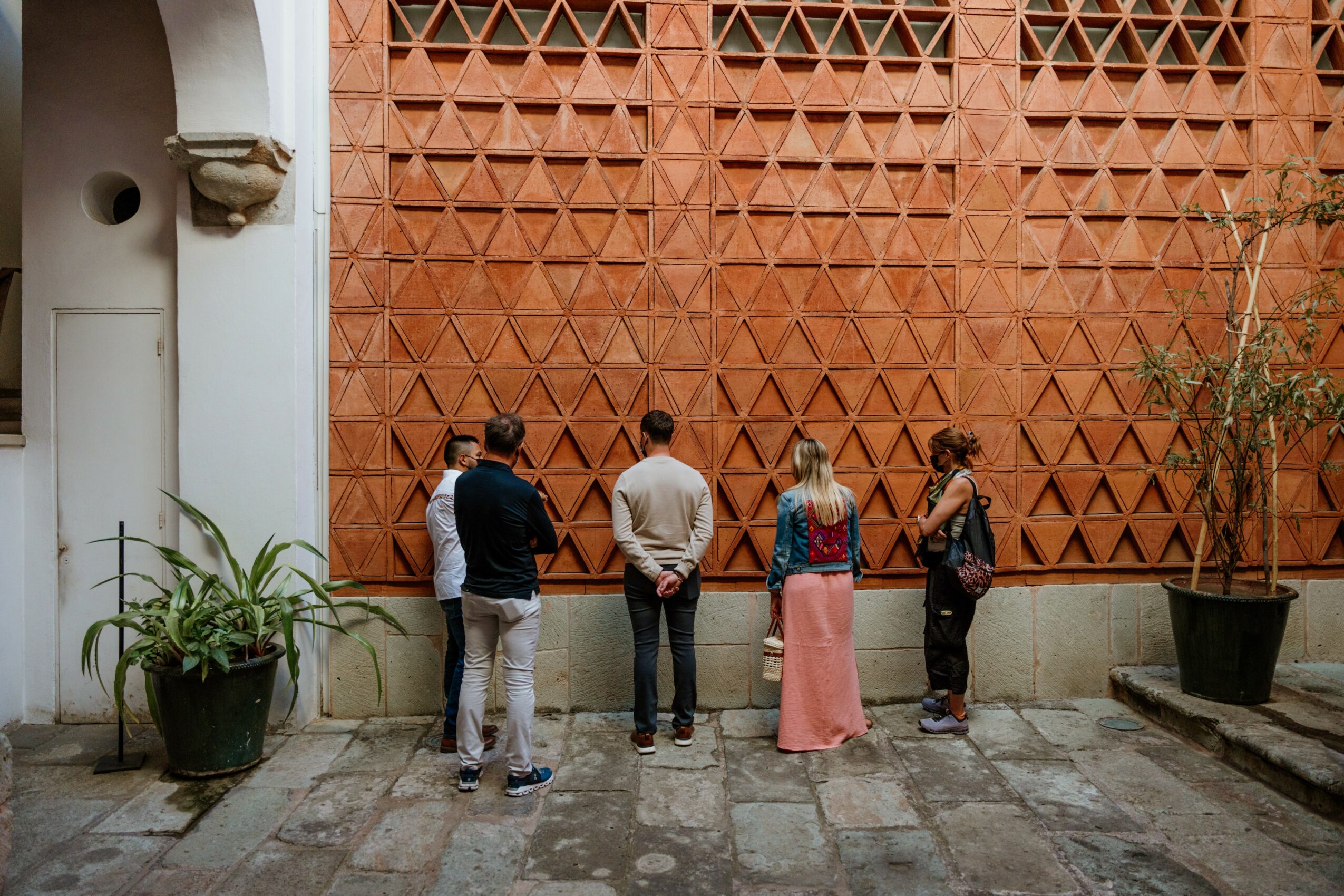 Four people stand in front of a decorative brick wall, observing it, with potted plants on either side.