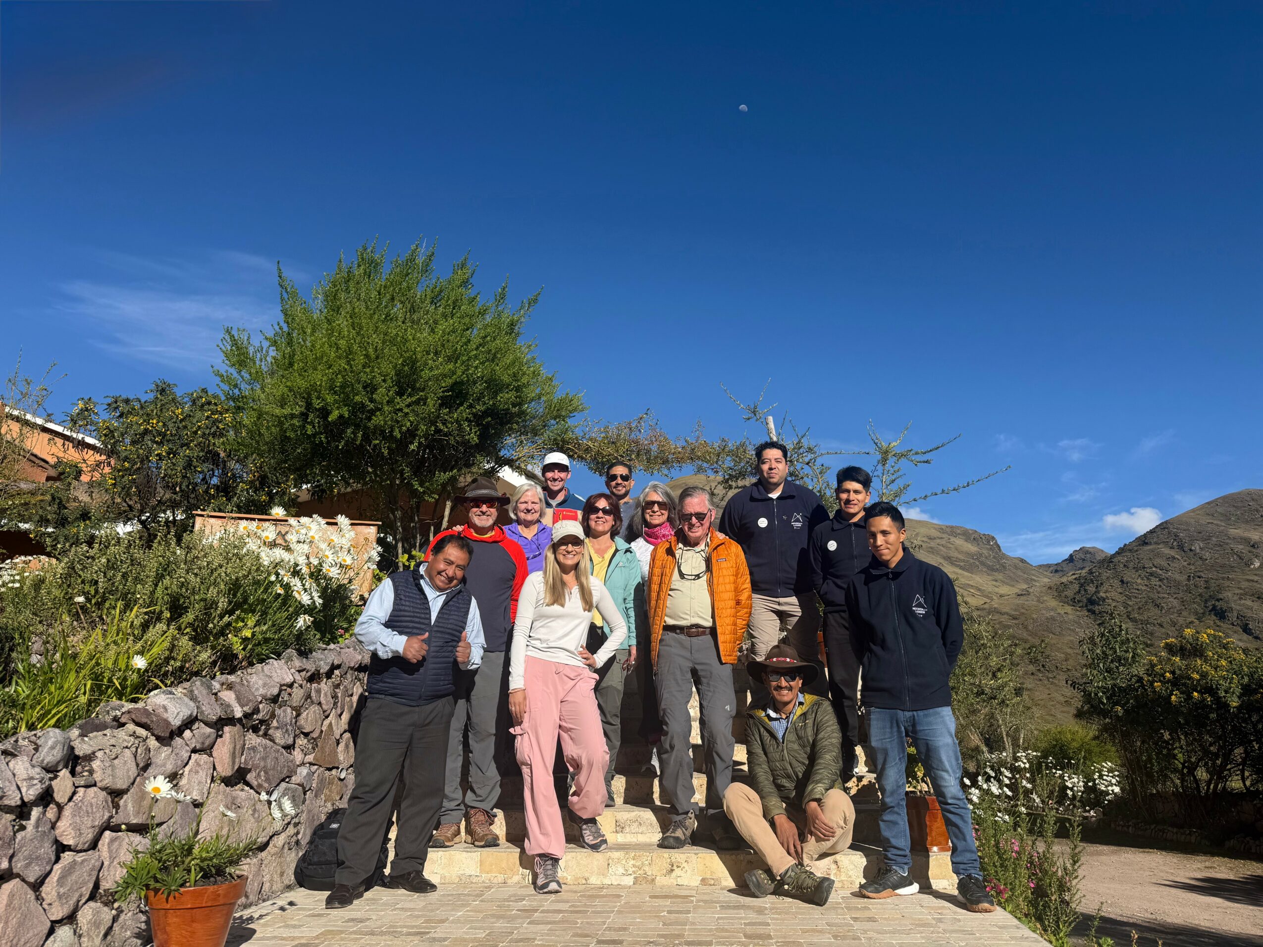 Group of ten people standing outdoors on a stone pathway with trees and mountains in the background under a blue sky.