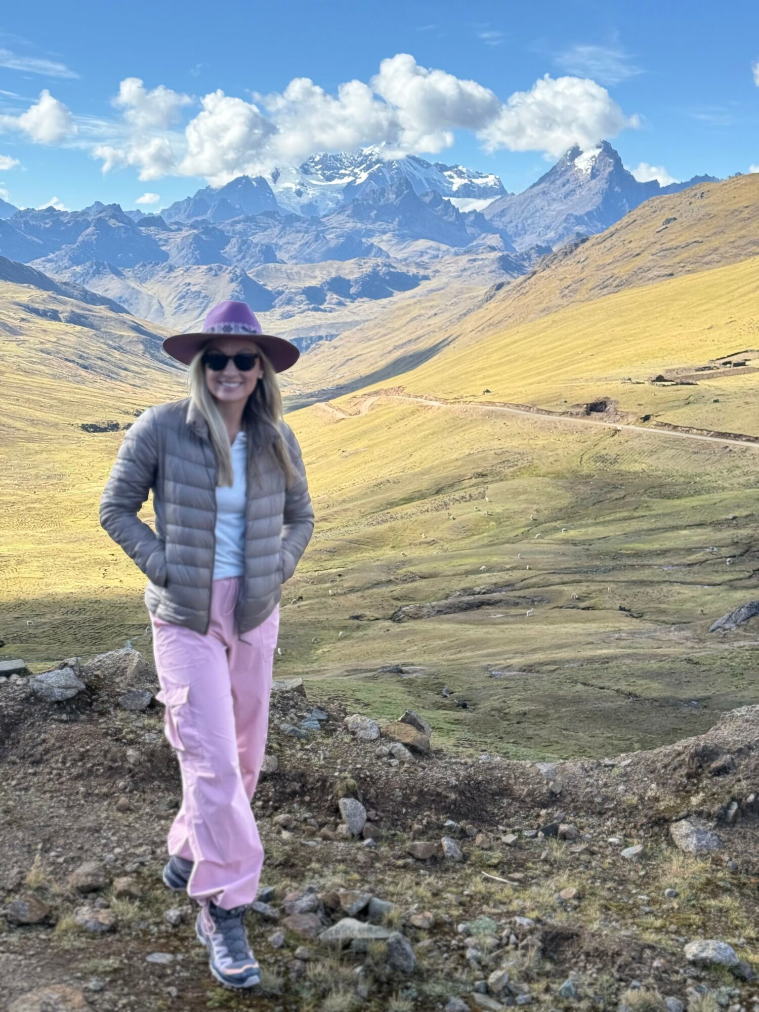 Woman standing outdoors in a mountainous landscape with grassy hills and snow-capped peaks, wearing a hat, sunglasses, and casual clothing.