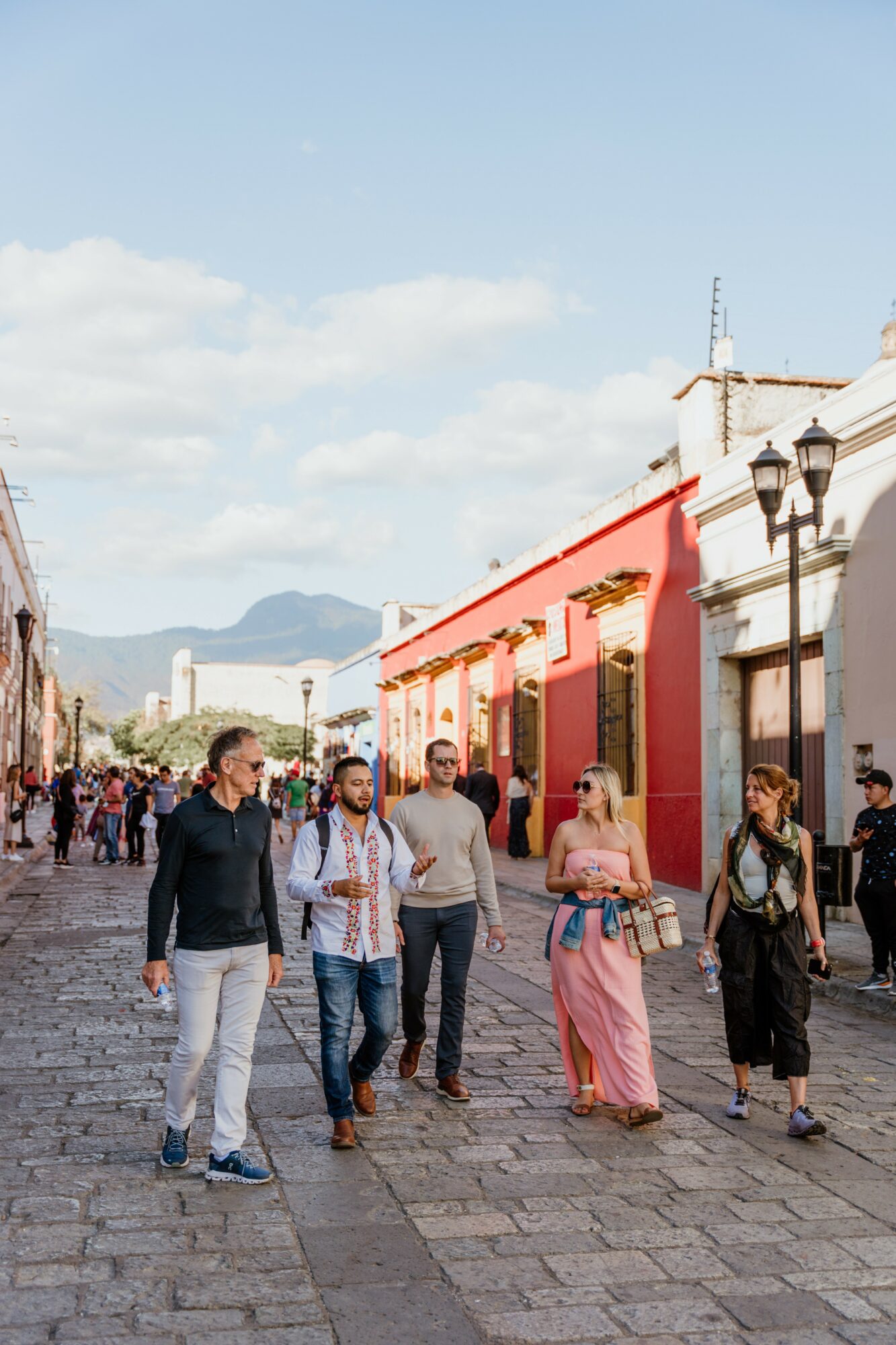 People walking on a cobblestone street with colorful buildings and a mountain in the background.