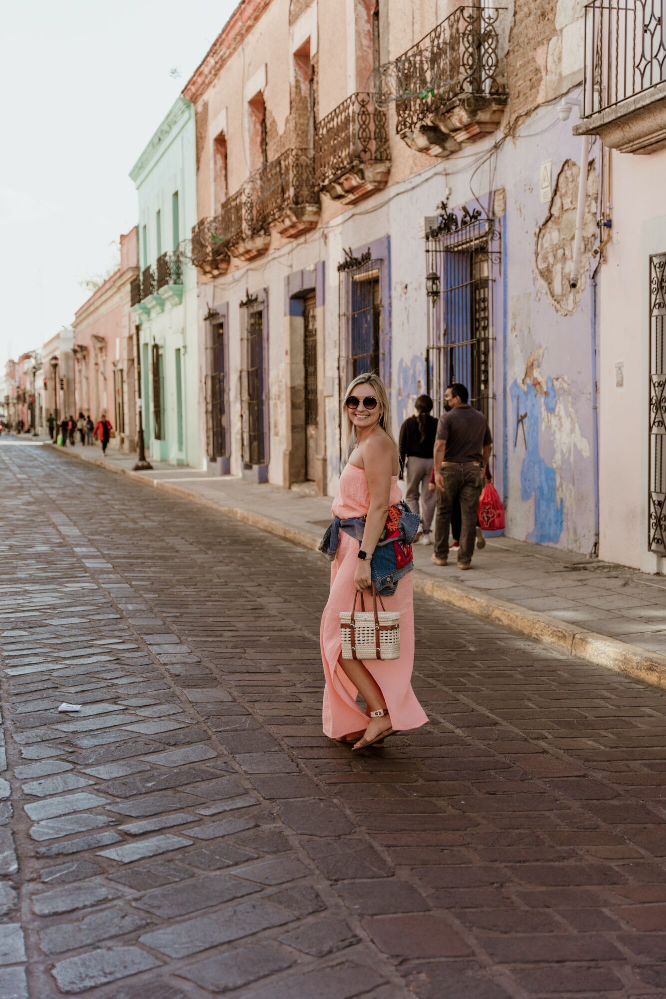 Woman in pink dress and sunglasses standing on cobblestone street with colorful old buildings behind her.