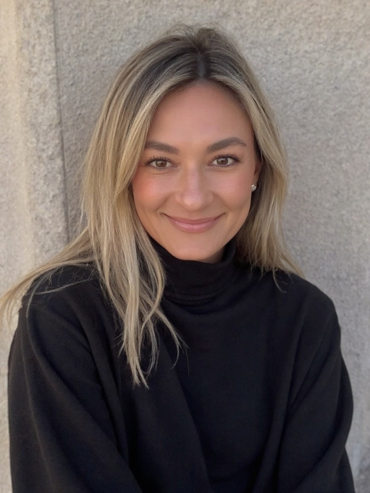 Young woman with blonde hair smiling, wearing a black turtleneck, standing against a textured wall.