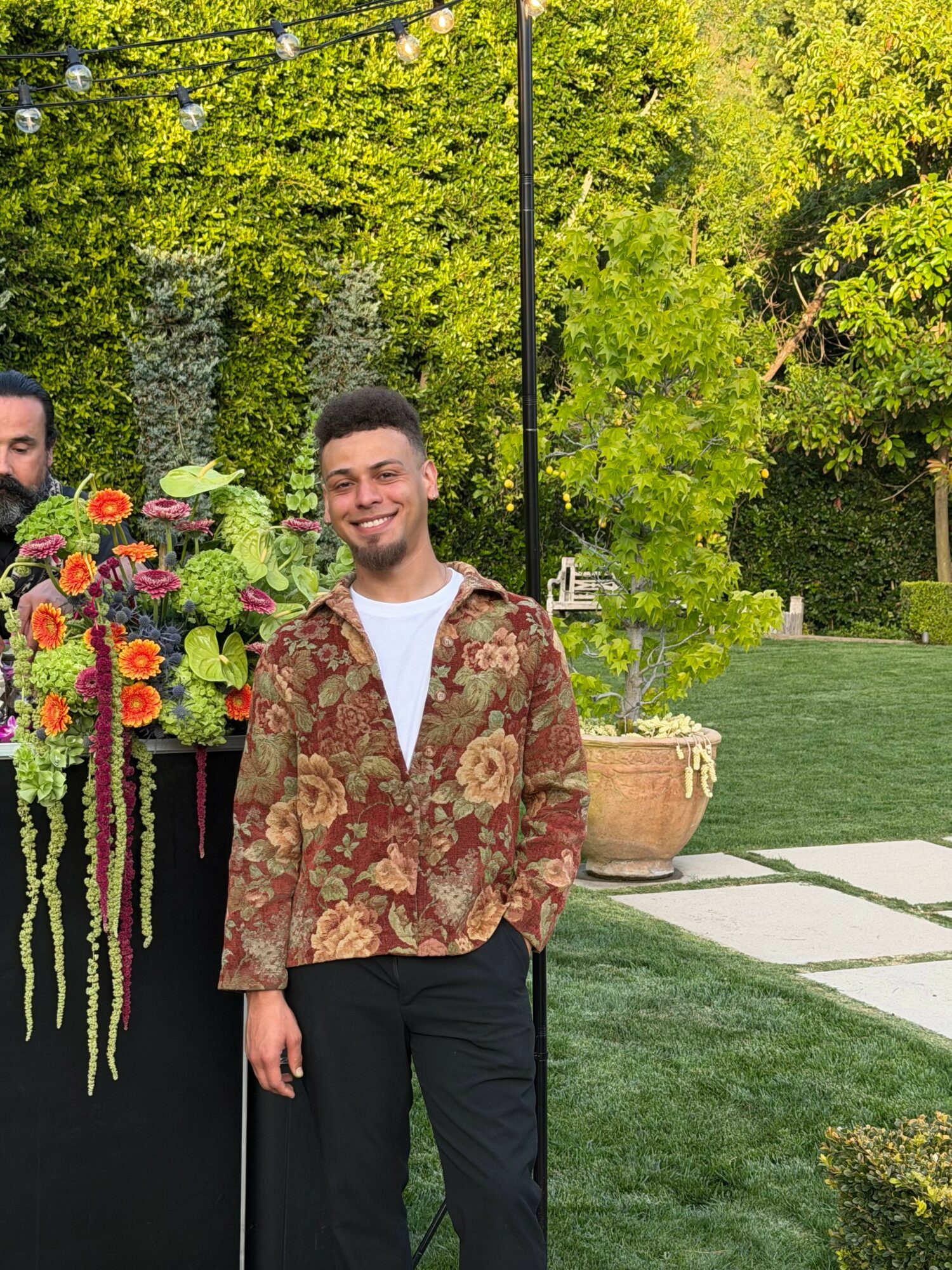Young man smiling outdoors, standing near colorful flower arrangement, green trees and lawn in background.