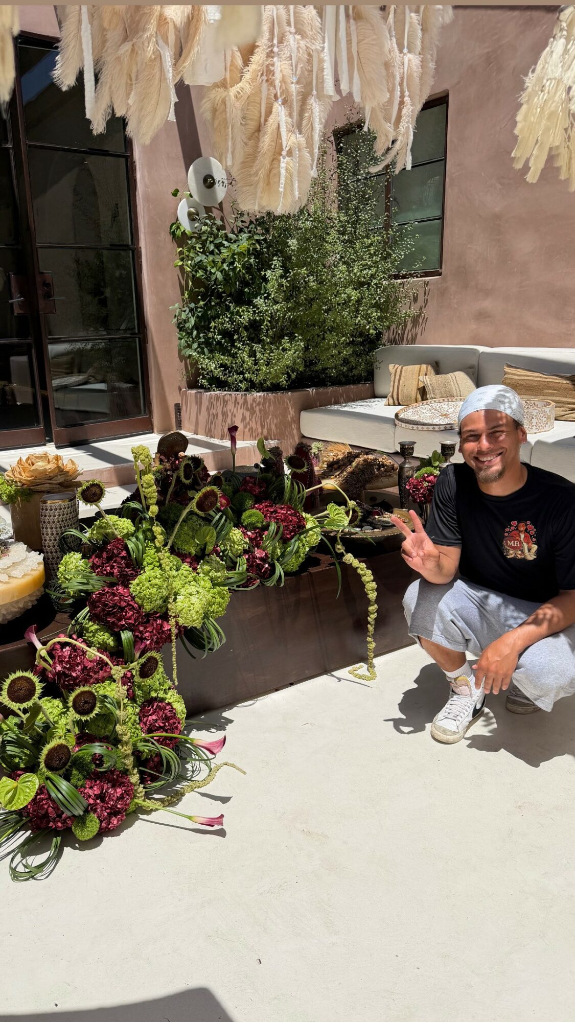 Person crouching next to colorful flower arrangements outdoors, with plants and a building in the background.