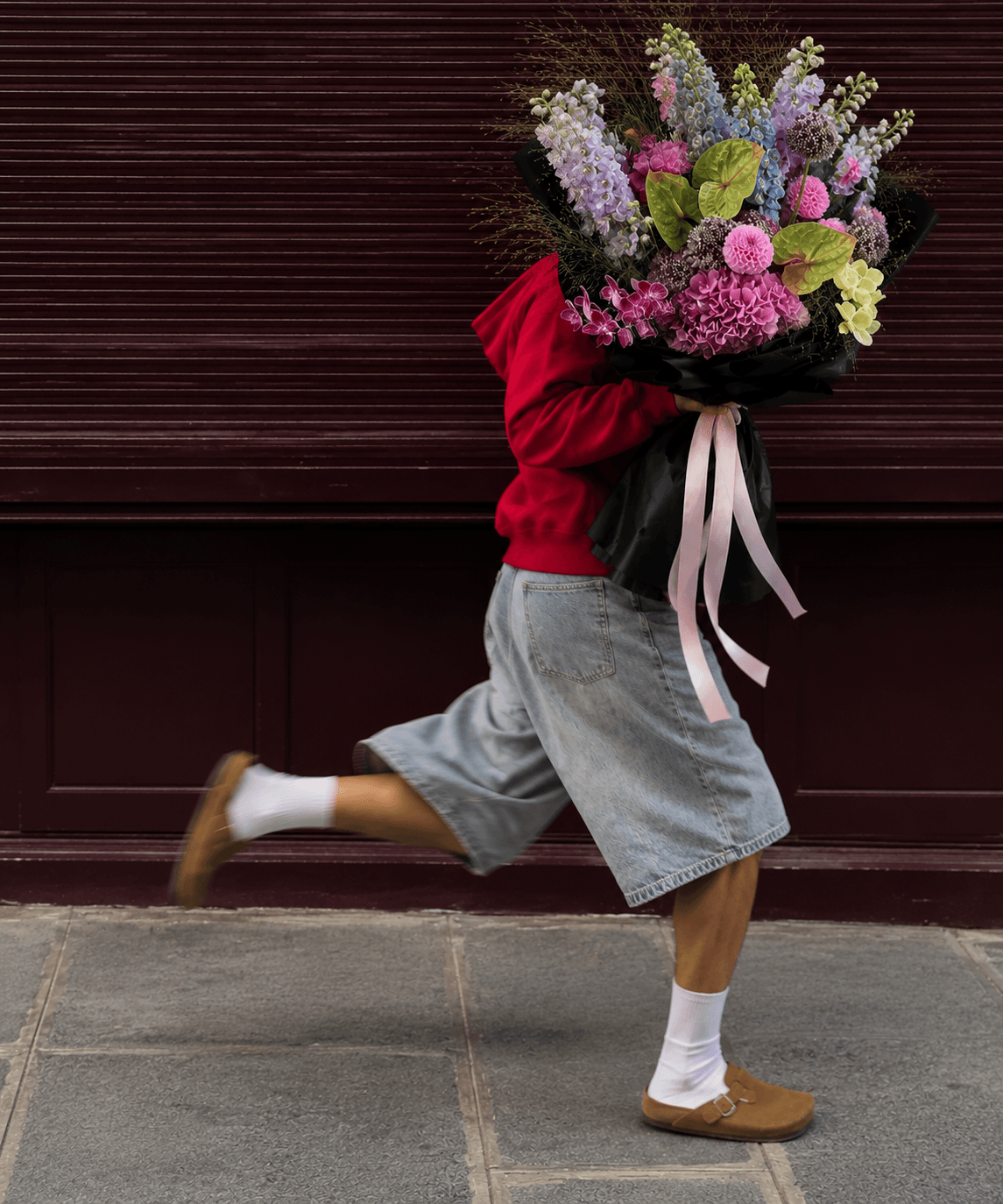 Child holding large bouquet of colorful flowers, wearing red sweater, gray shorts, white socks, and brown shoes, mid-stride.