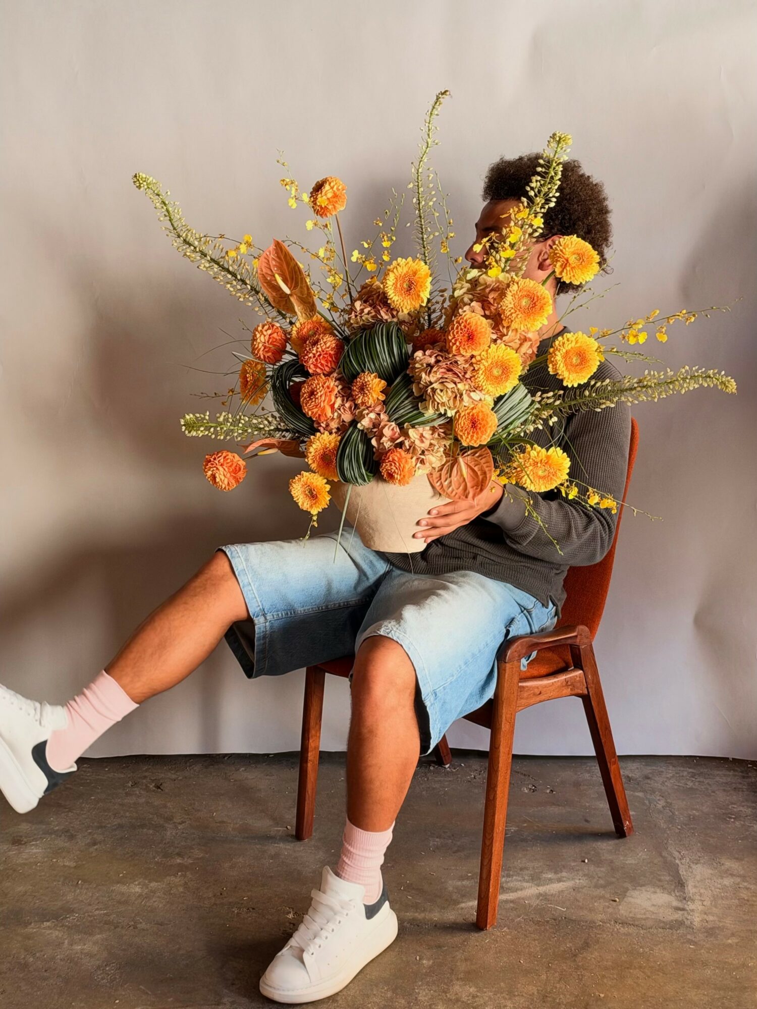 Child sitting on a wooden chair holding a large bouquet of orange and yellow flowers, with a neutral background.