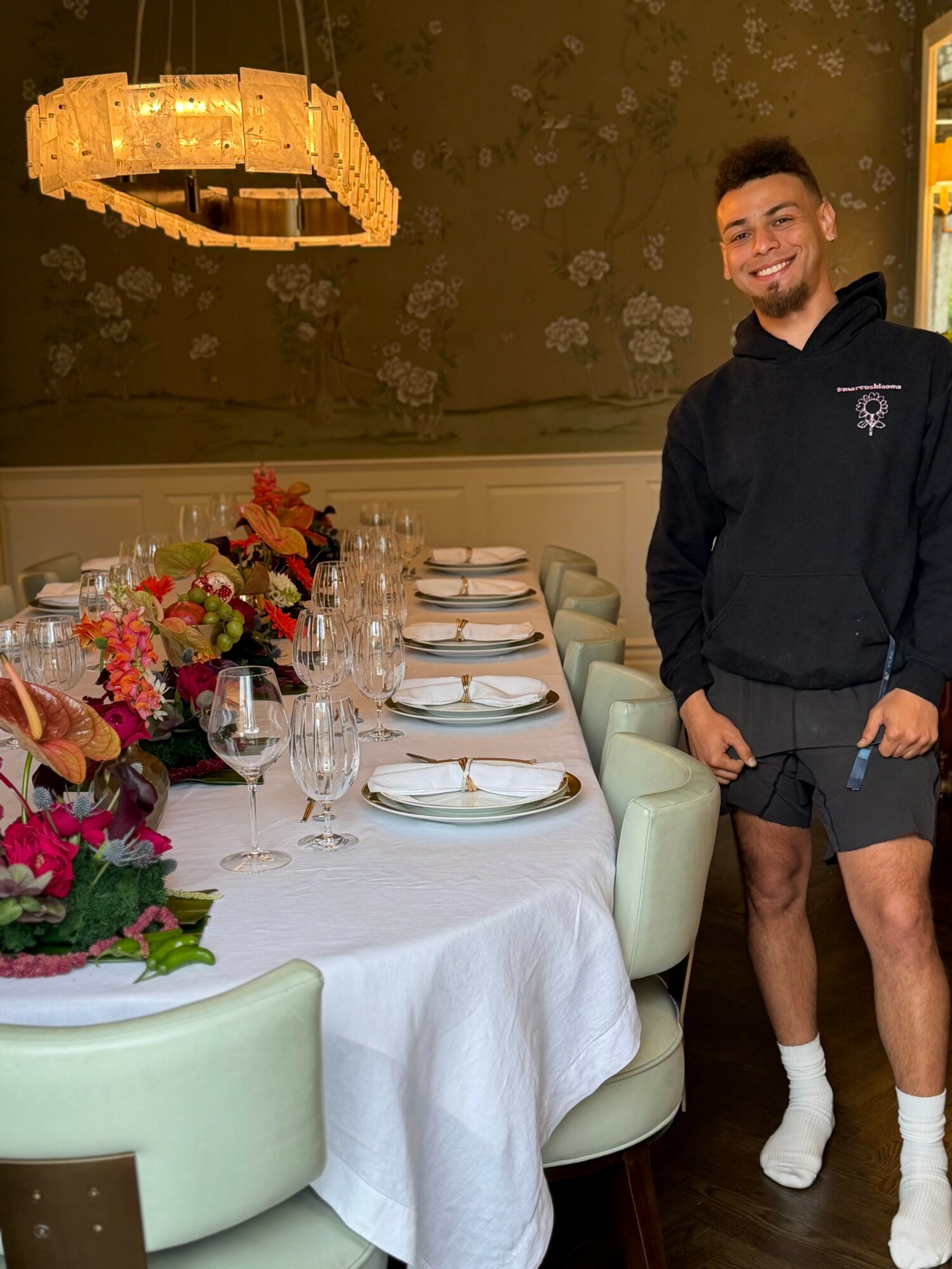 Young man standing next to a dining table with floral arrangements, set with plates and glasses, in a decorated room.