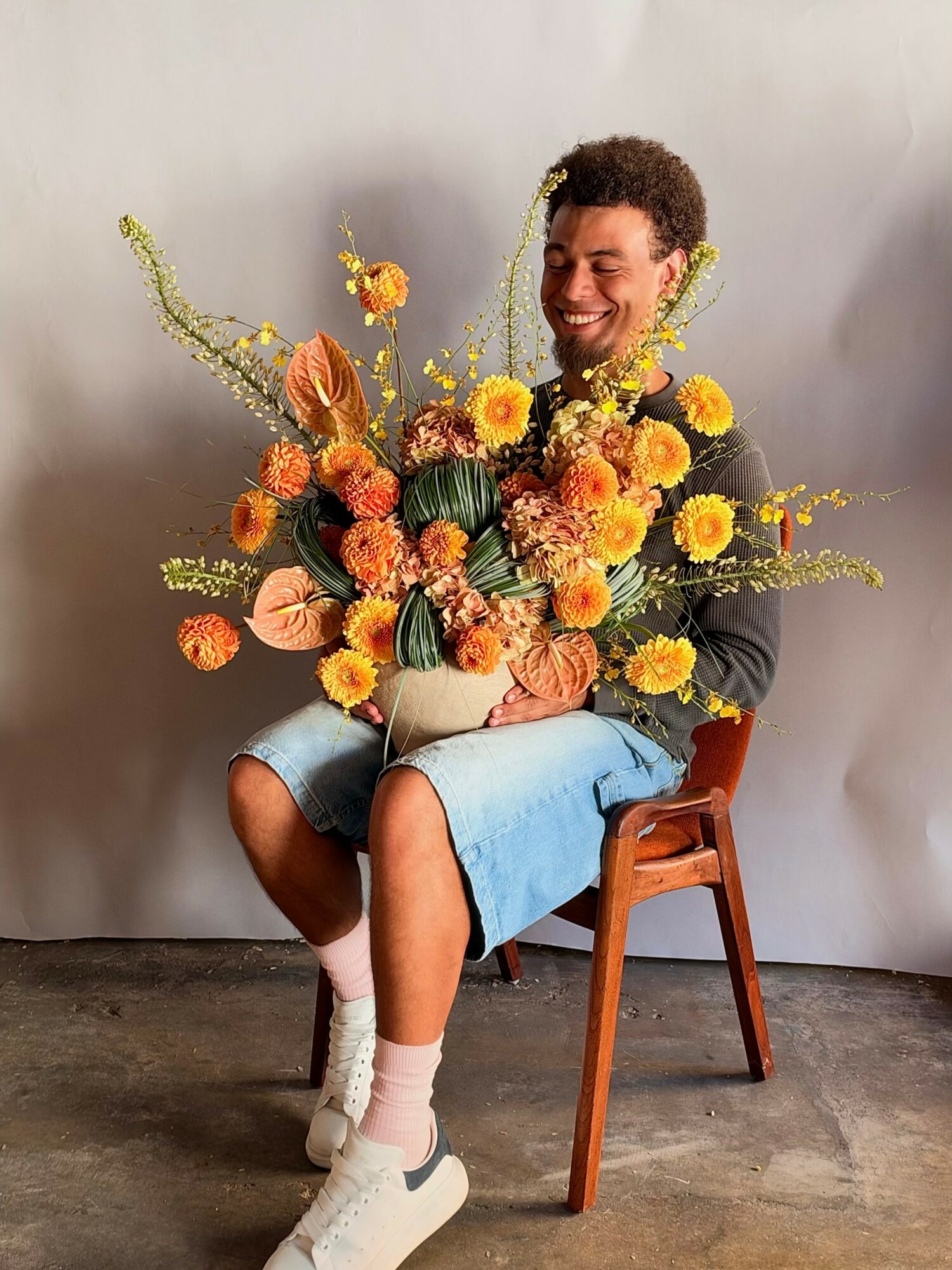 Young man sitting on a wooden chair holding a large colorful flower arrangement, smiling, against a plain background.
