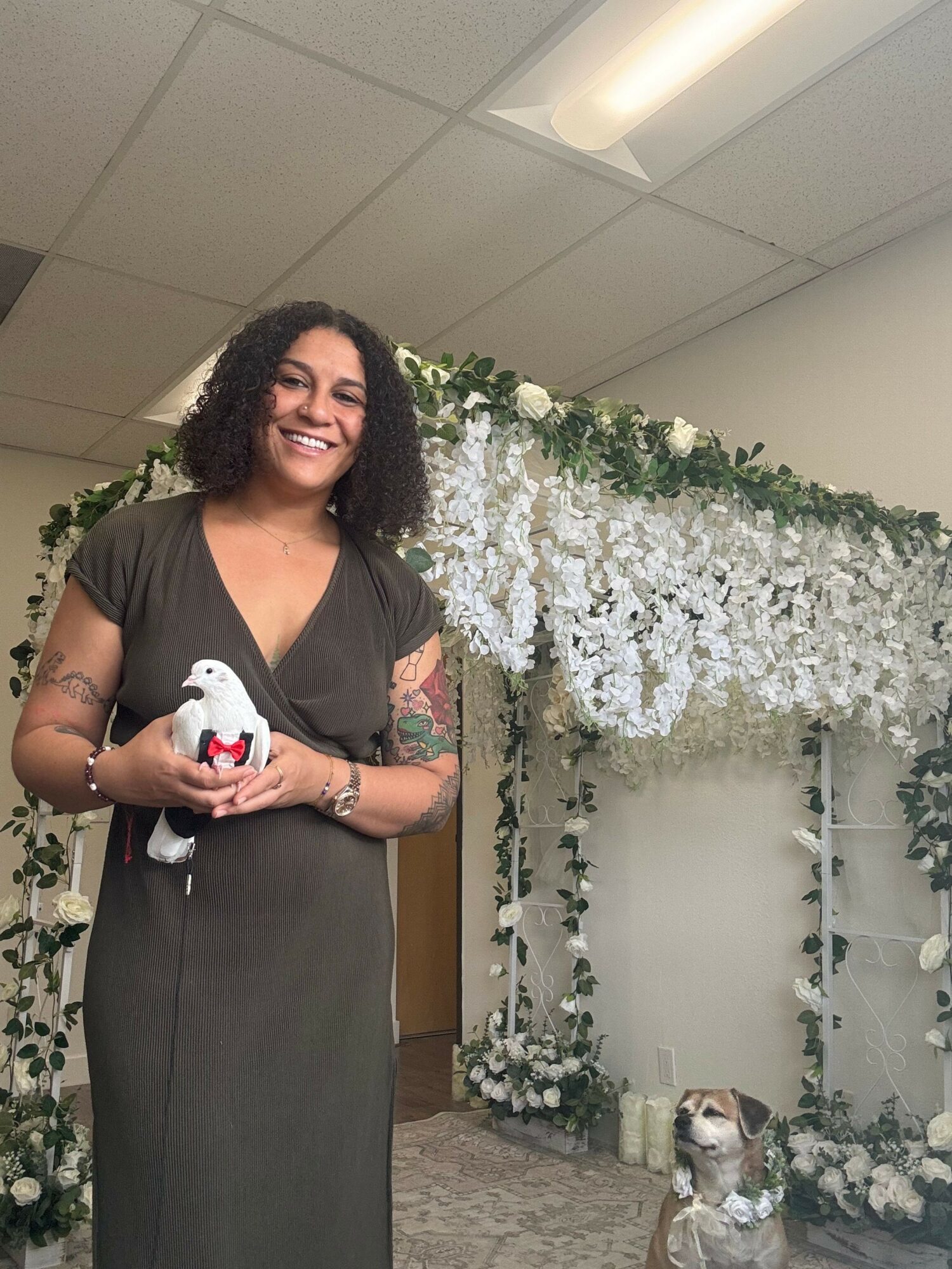 Woman in black dress holding a stuffed animal, smiling, standing in front of floral backdrop with white flowers and greenery.