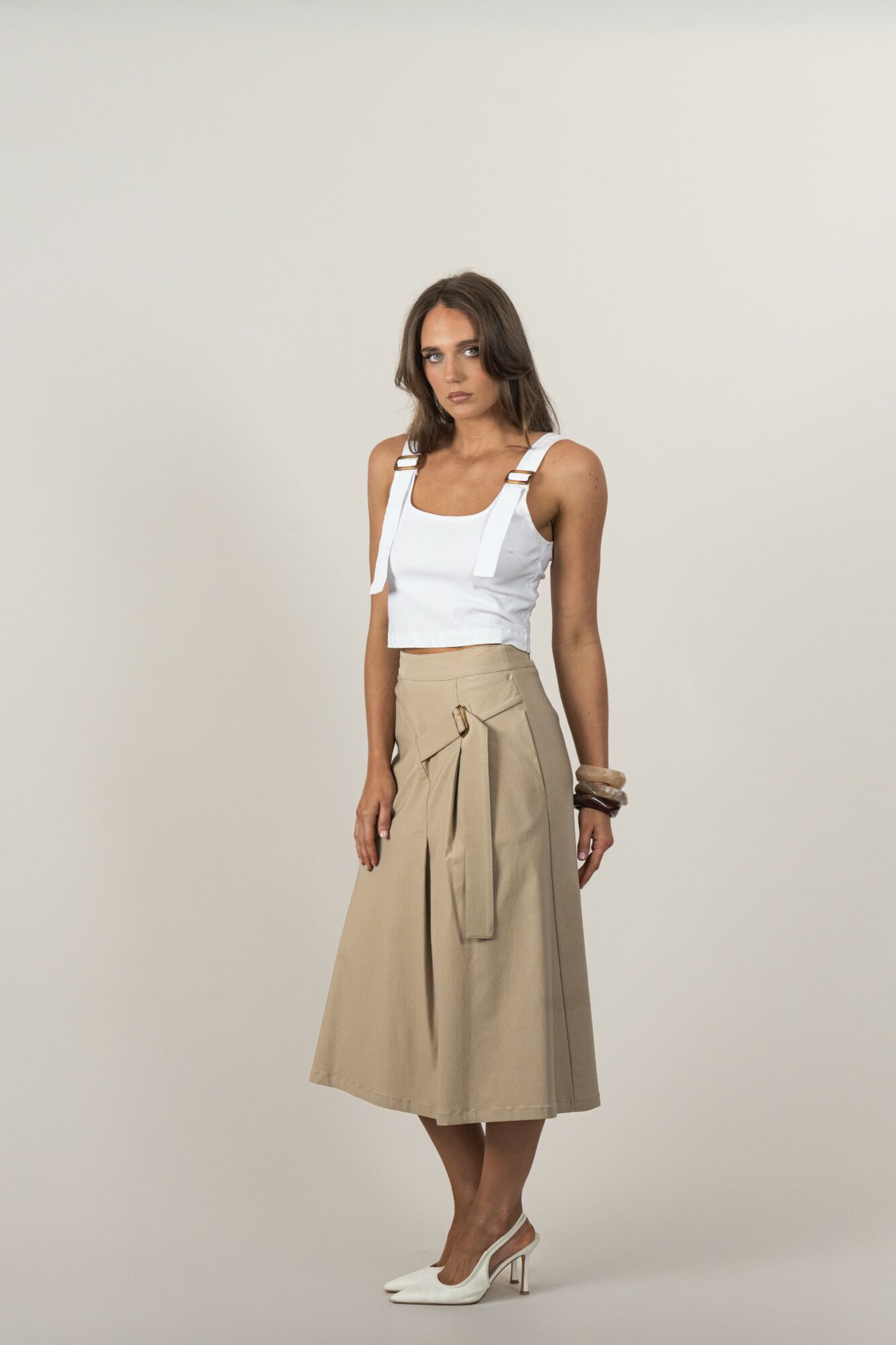 Woman standing against a plain background, wearing a white top, beige skirt, and high heels, looking at the camera.