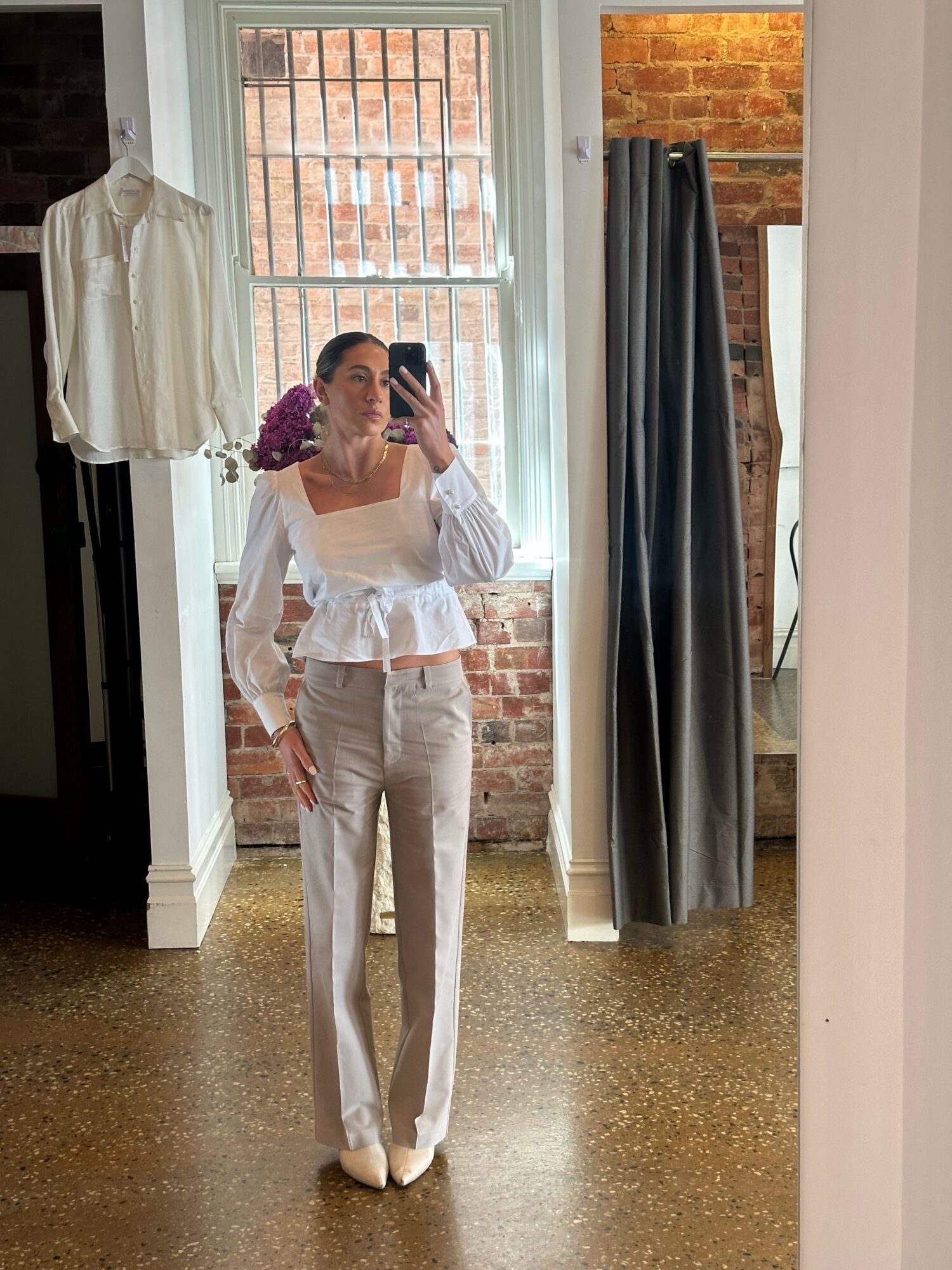 Person in white outfit holding a bouquet of purple flowers, standing in a room with brick wall and window, mirror, and curtains.