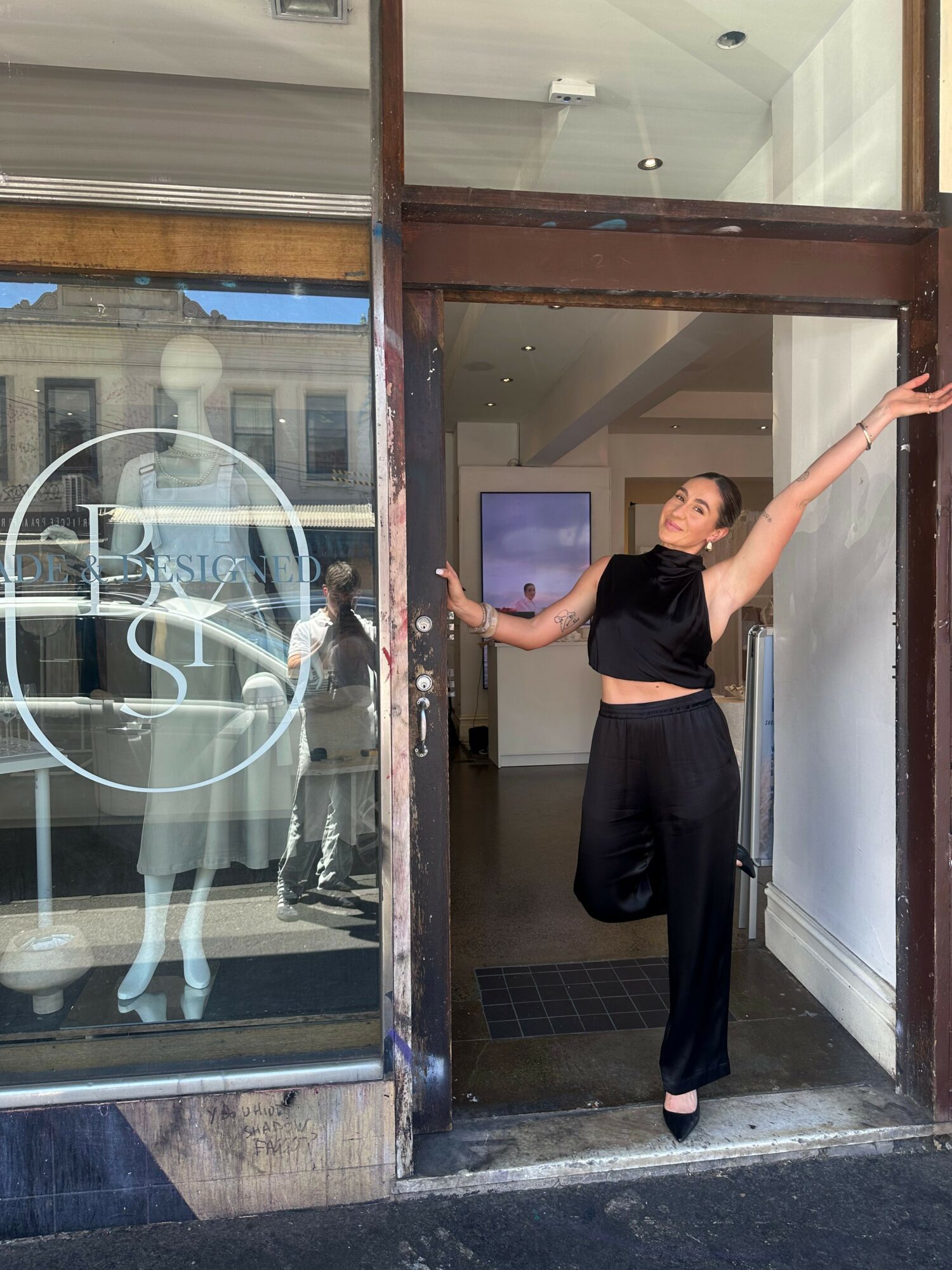 Person standing in doorway, one arm raised, wearing black outfit, reflected in glass window, urban street scene behind.