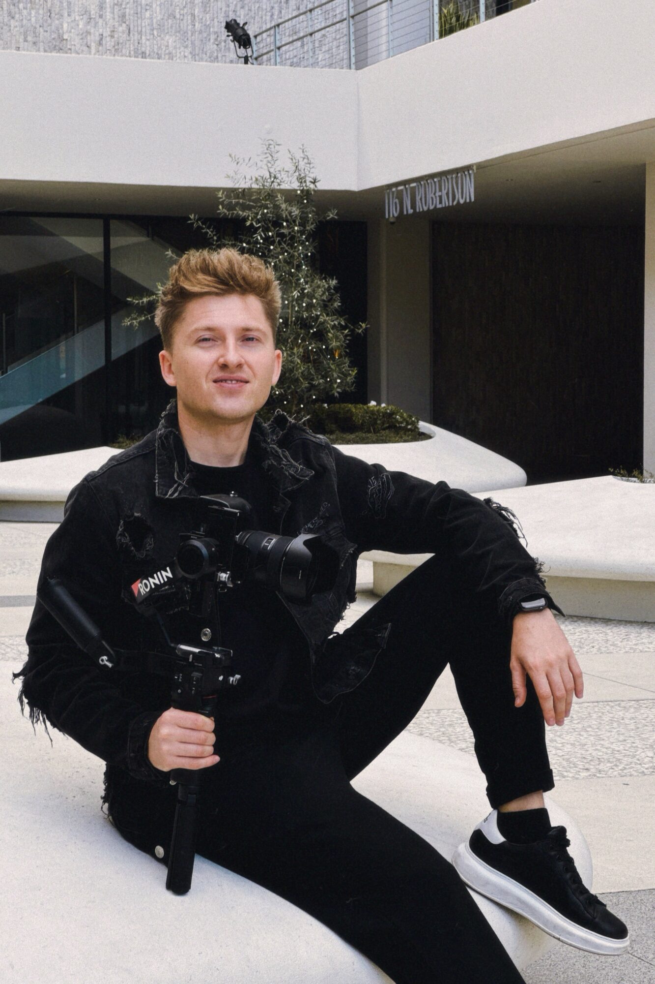 Young man with short hair sitting outdoors, wearing black clothing and sneakers, in front of modern building.