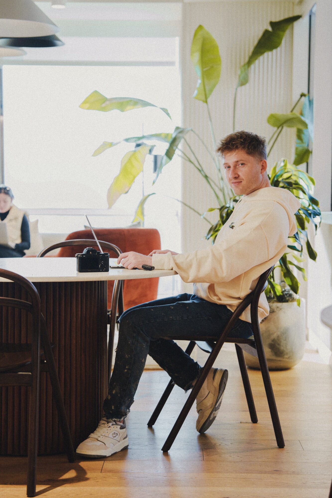 Young man sitting at a table in a bright room with large plants and natural light, looking at the camera.