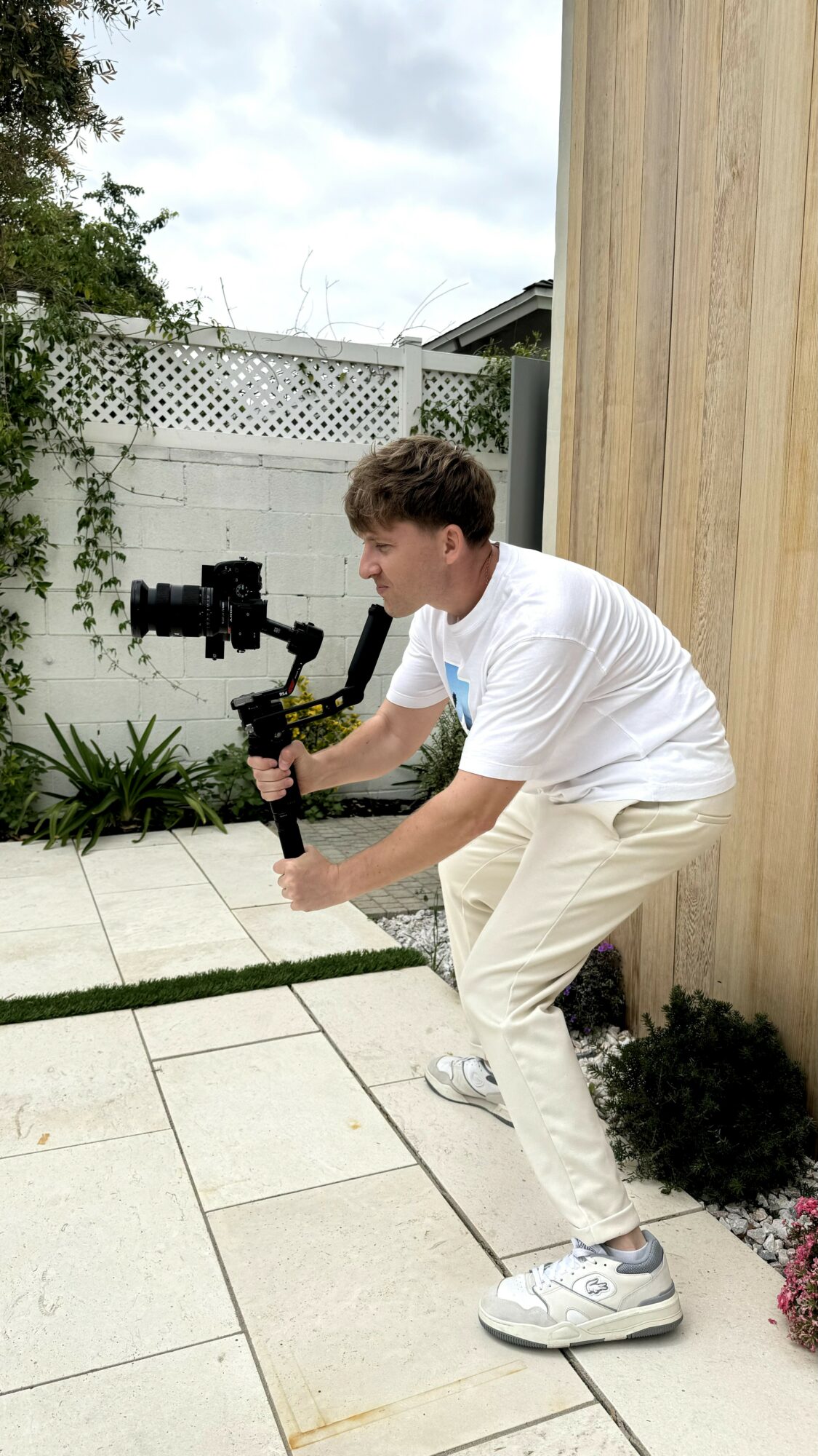 Young person in white shirt and beige pants holding a camera stabilizer outdoors near a wooden wall, with plants and a white fence in background.