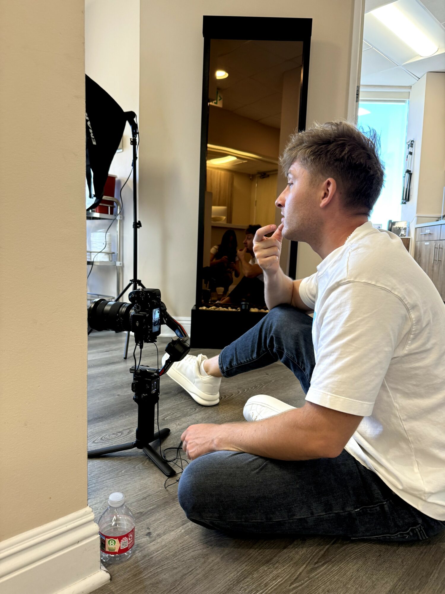 Person sitting on the floor with a camera setup, drinking from a cup, with a mirror and a water bottle nearby.