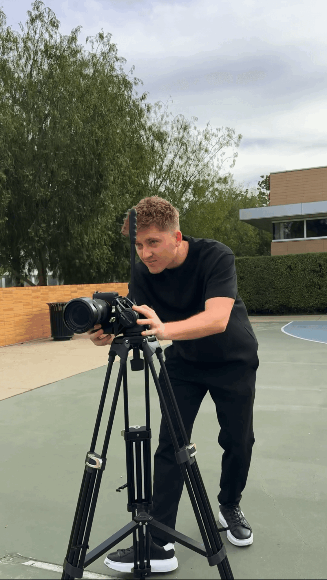 Young man adjusting camera on tripod outdoors, trees and building in background, overcast sky, wearing black shirt and pants.
