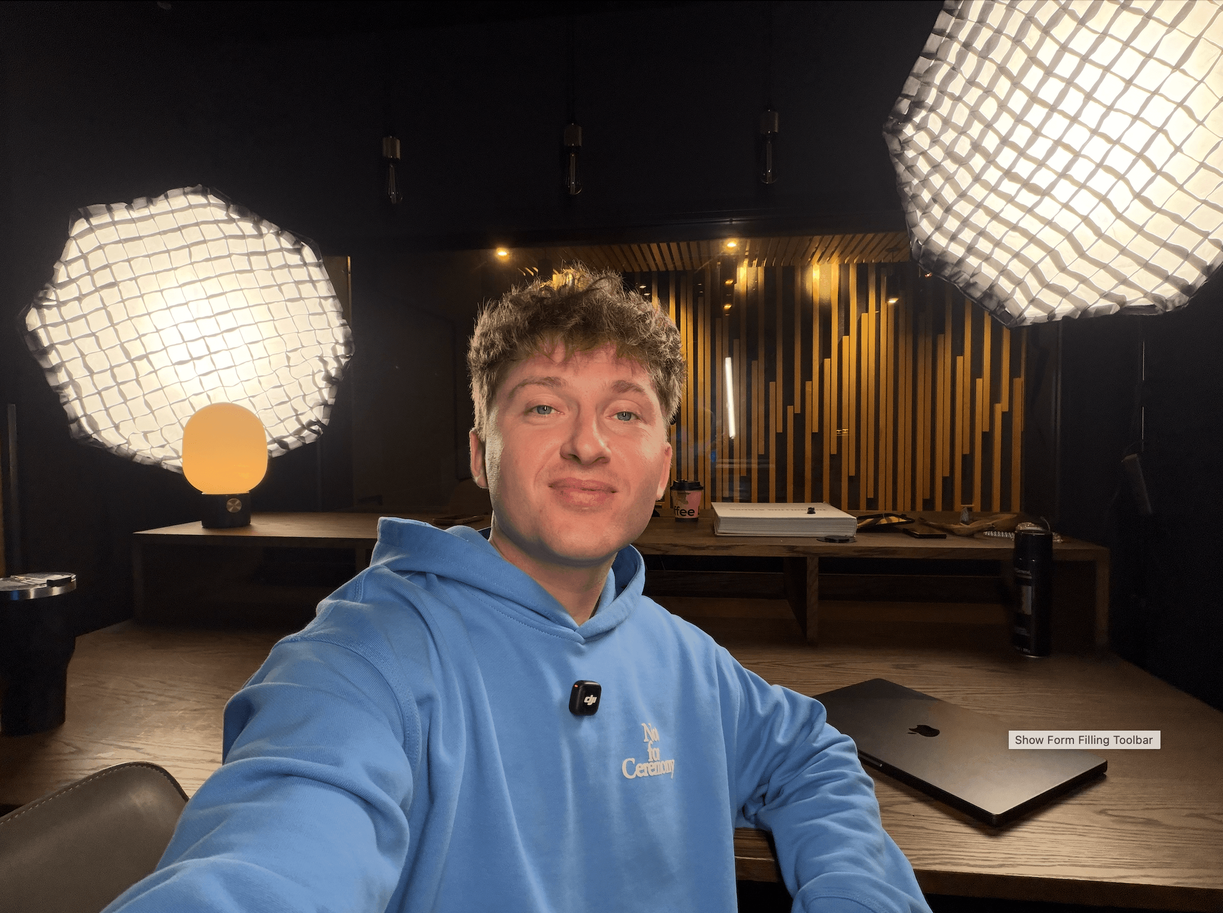 Young man with curly hair in a blue hoodie taking a selfie in a room with large, white, textured pendant lights and a dark background.