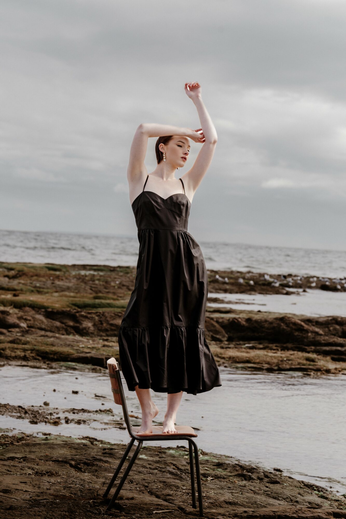 Woman standing on a chair on a rocky beach with cloudy sky and ocean in background, arms raised above head.