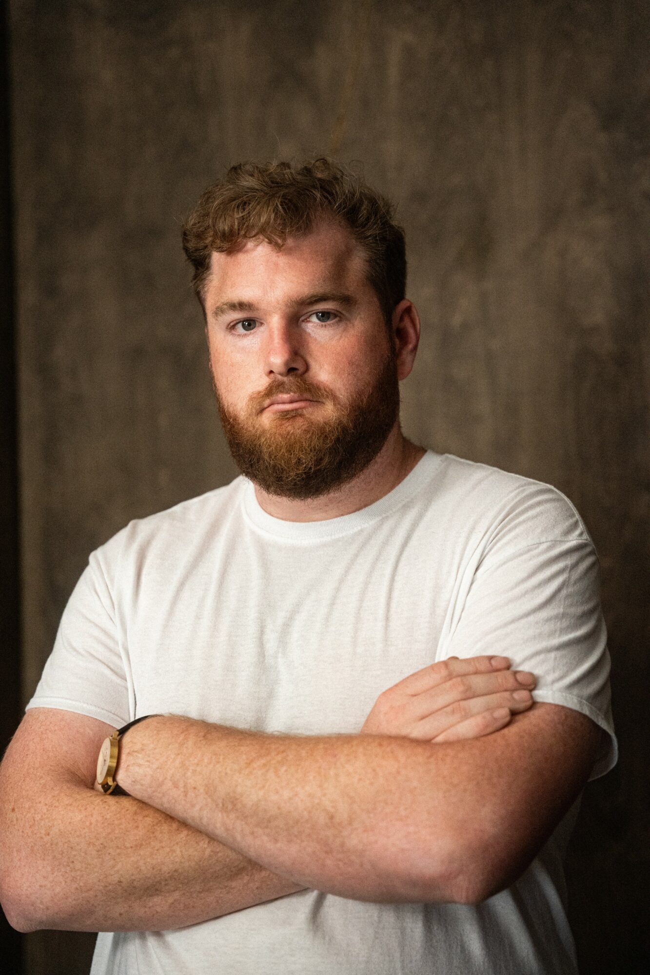 Man with curly hair and beard wearing a white t-shirt, arms crossed, against a textured brown background.