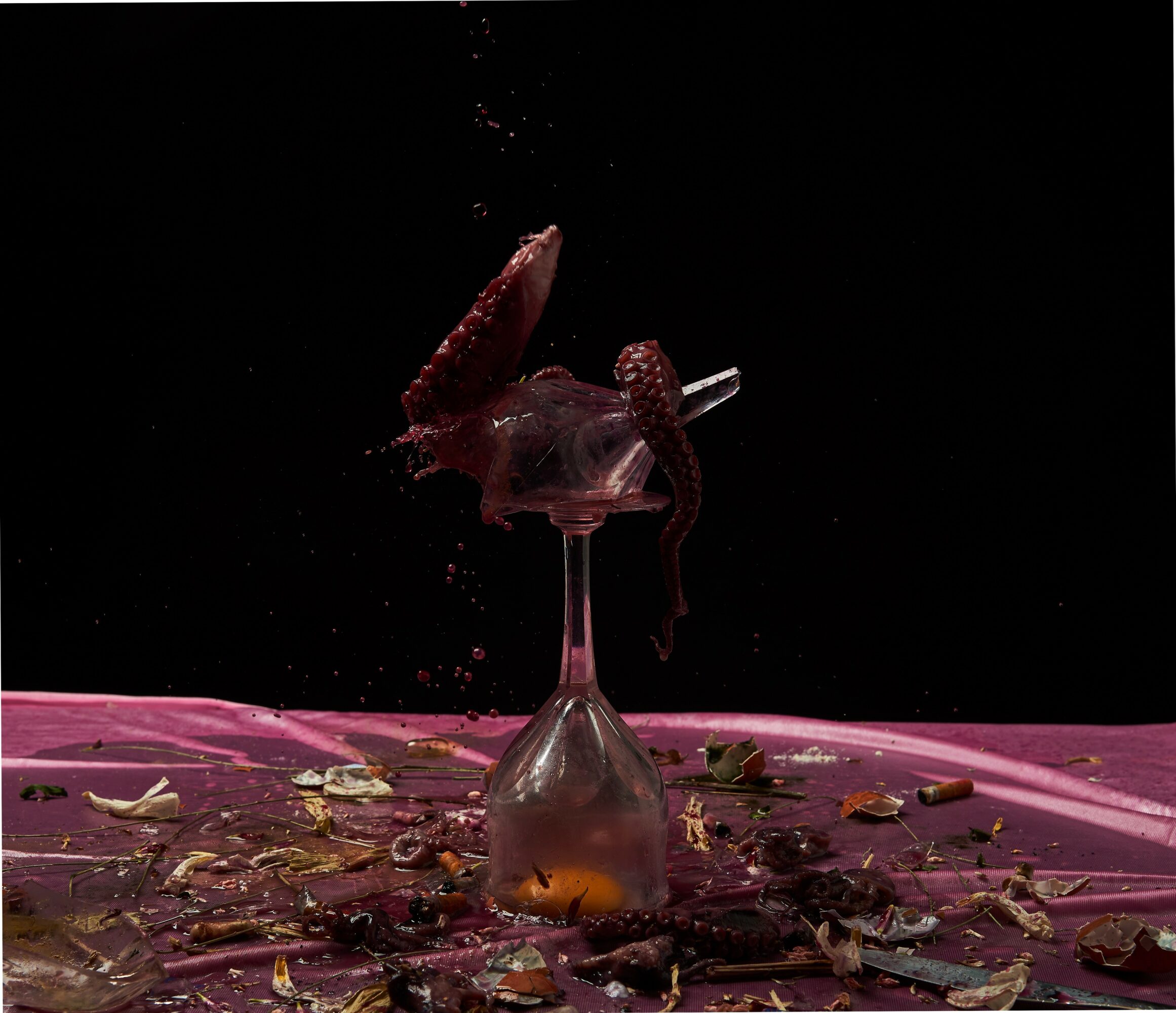 A glass with strawberries and liquid splashing out, on a pink surface with scattered debris, against a black background.