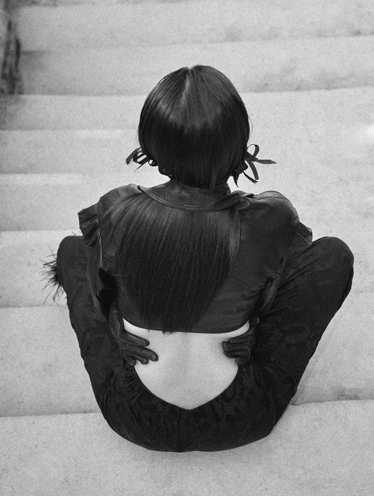 Person sitting on stairs with back facing camera, legs crossed, wearing dark clothing, hair tied with ribbons.